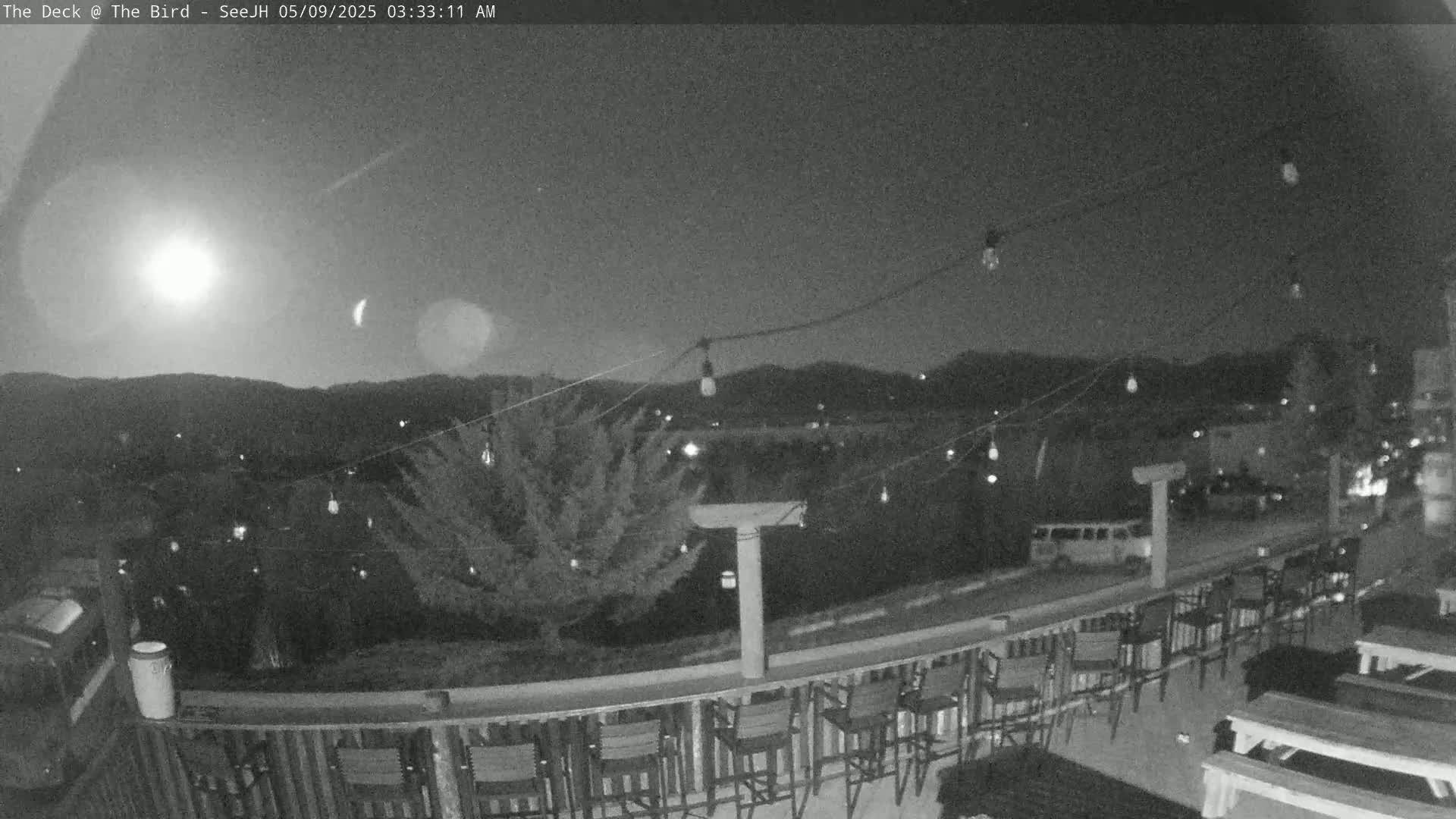 A nighttime view from a deck shows a string of lights above an outdoor bar area overlooking a moonlit landscape of hills and distant buildings.