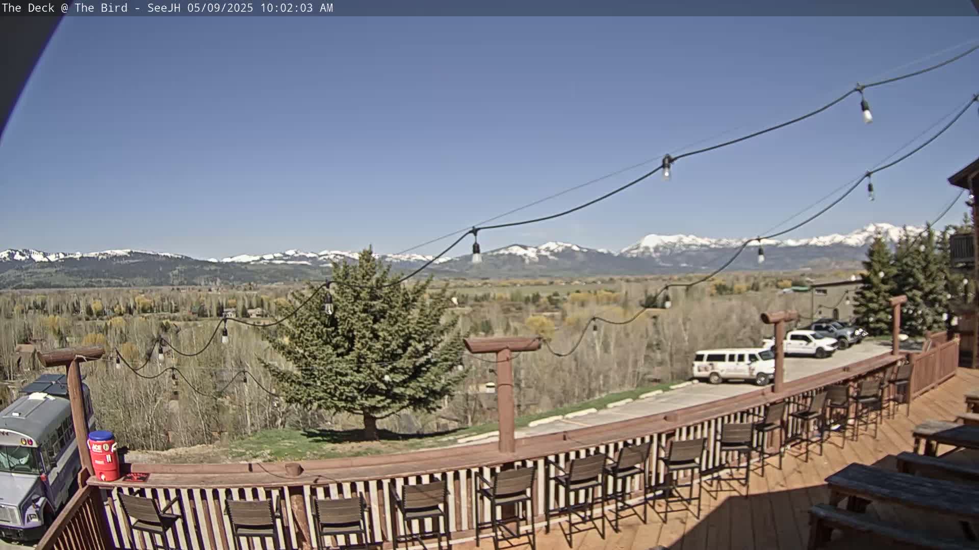 A deck with bar stools and picnic tables overlooks a valley with snow-capped mountains in the background under a clear blue sky.