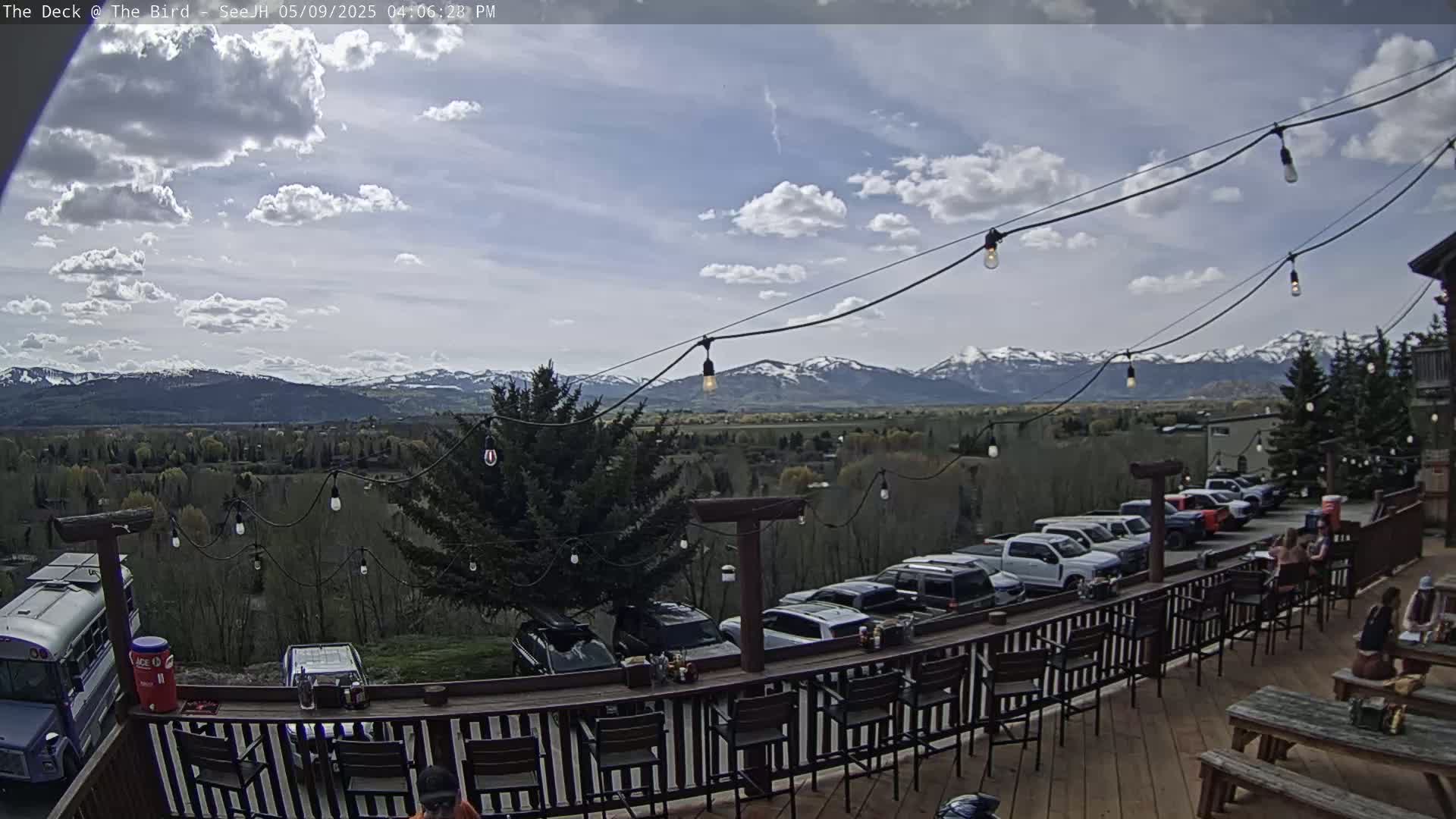 A deck with string lights overlooks a parking lot and a valley with snow-capped mountains in the distance under a partly cloudy sky.