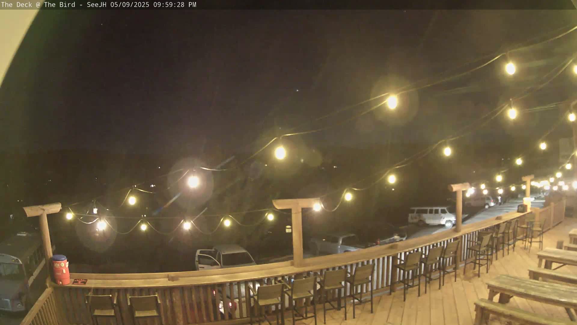 A nighttime view of an outdoor deck with string lights, wooden furniture, and a railing overlooking a parking lot.
