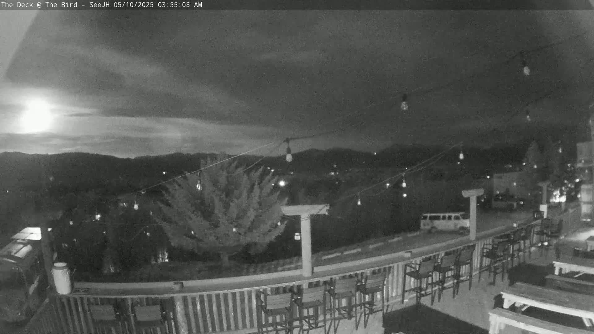 A nighttime view of an outdoor deck with string lights, empty bar stools, and picnic tables, overlooking a town nestled in hills under a partly cloudy sky.