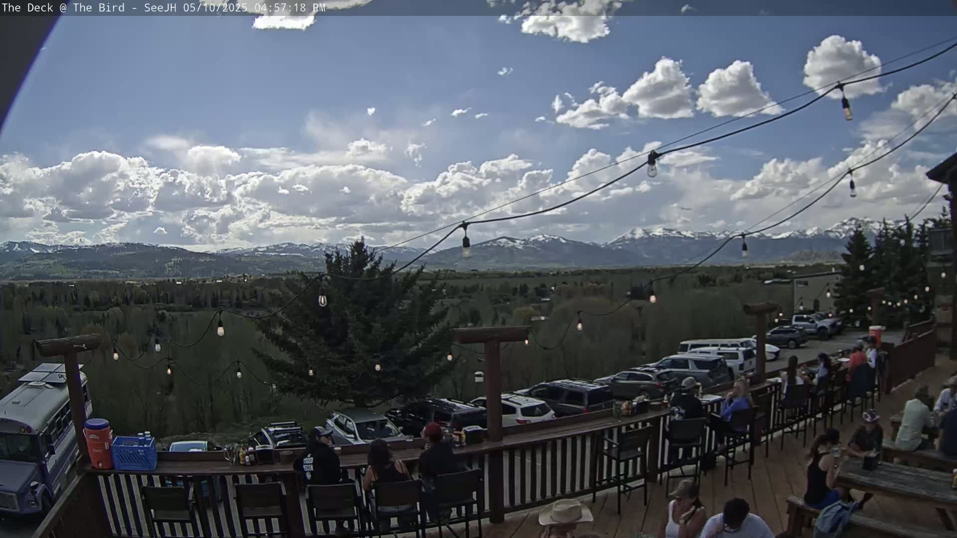 An outdoor patio with people seated at tables, overlooking a valley and snow-capped mountains under a partly cloudy sky.