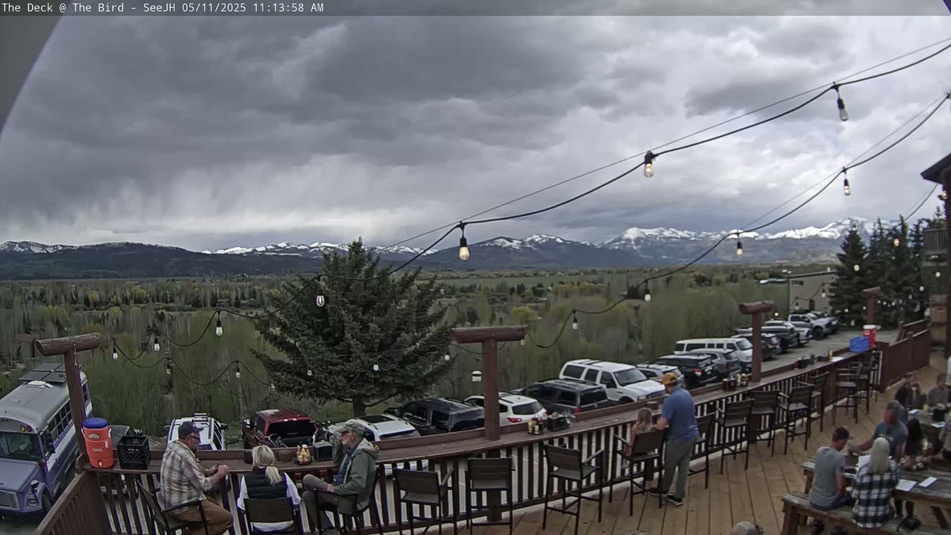 An outdoor deck with people seated at tables and a bar overlooks a valley and snow-capped mountains under a cloudy sky.