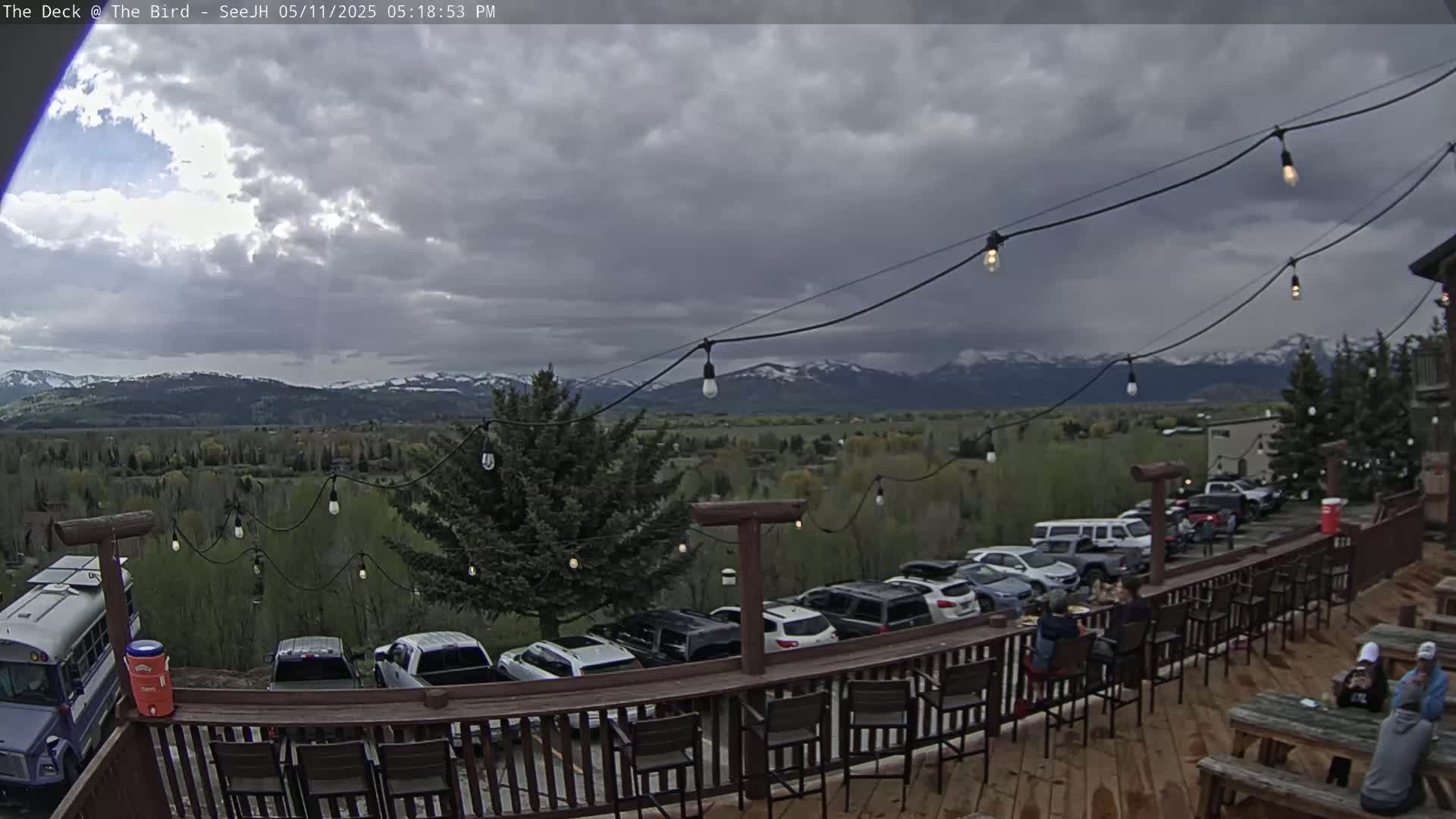 An outdoor deck with string lights overlooks a parking lot and a valley with snow-capped mountains under a cloudy sky.