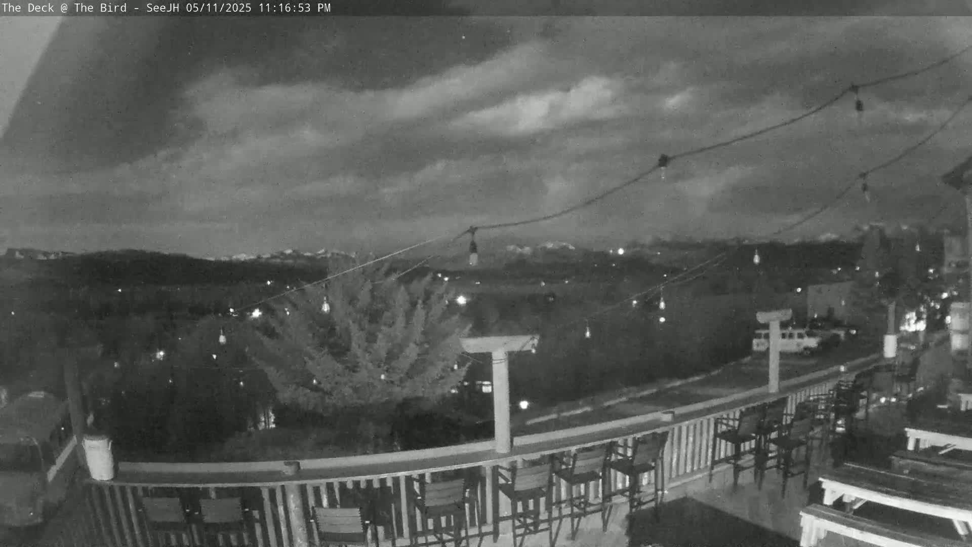 An outdoor deck with string lights and empty chairs overlooks a dimly lit town and distant mountains under a cloudy night sky.