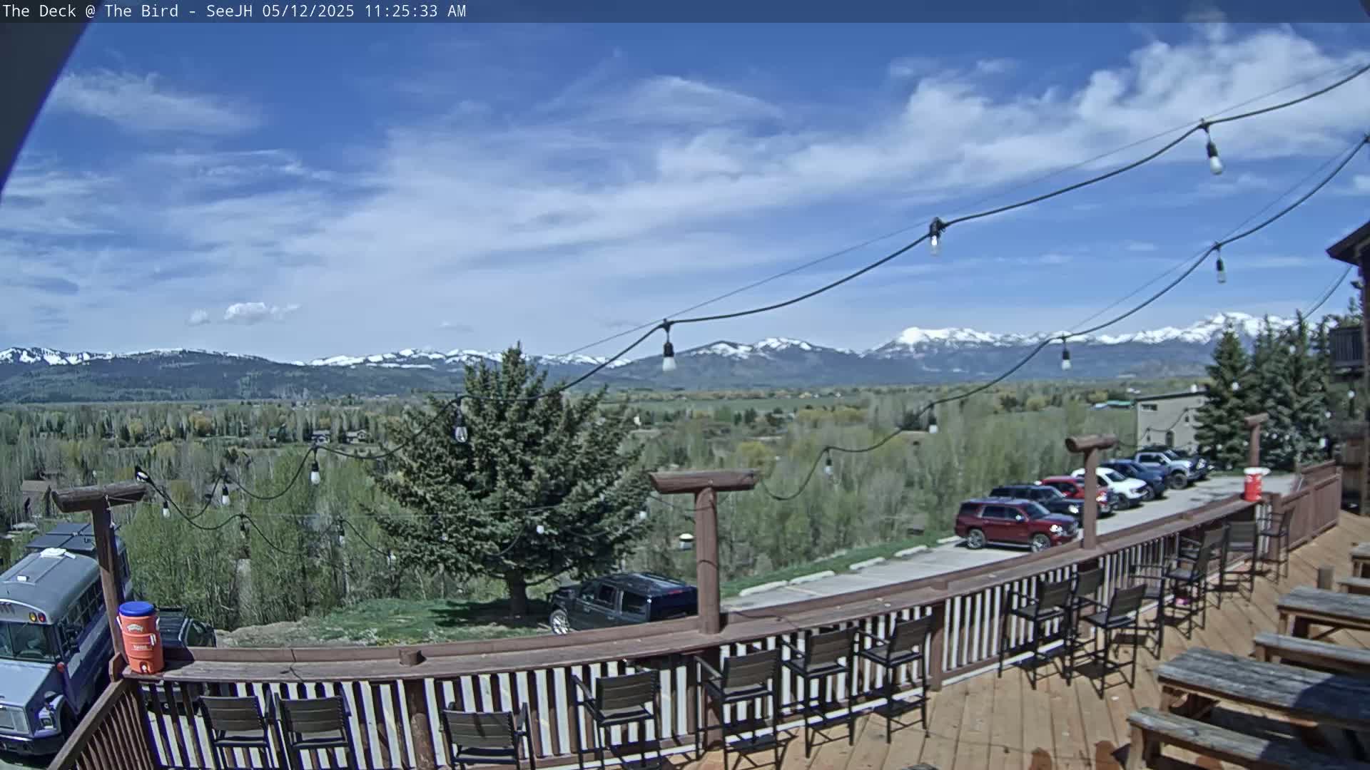 A deck with a wooden railing and outdoor seating overlooks a valley with snow-capped mountains in the distance under a partly cloudy sky.