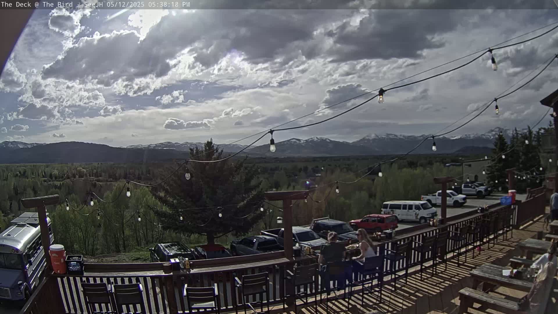 A partly cloudy outdoor patio scene overlooks a valley with mountains in the distance, featuring string lights, parked vehicles, and people seated at tables.