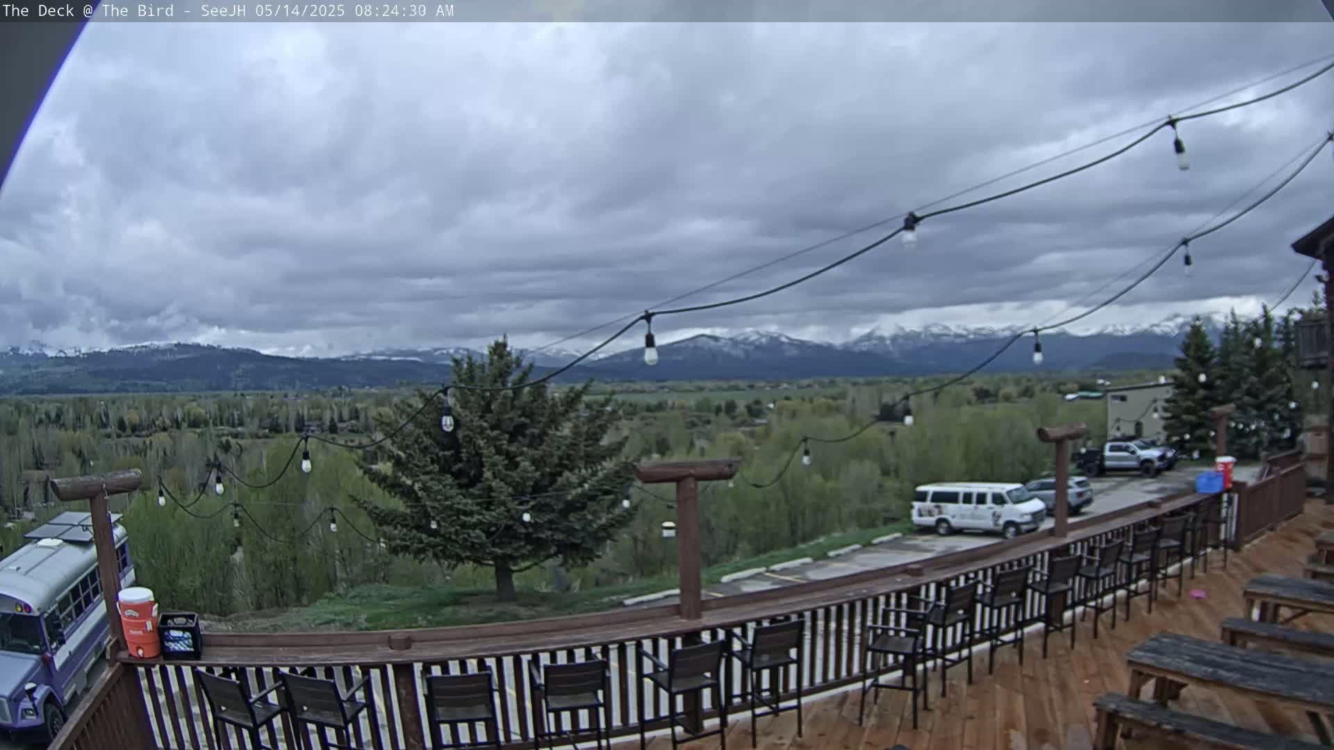 A deck with a wooden railing and string lights overlooks a valley with snow-capped mountains in the distance under a cloudy sky.
