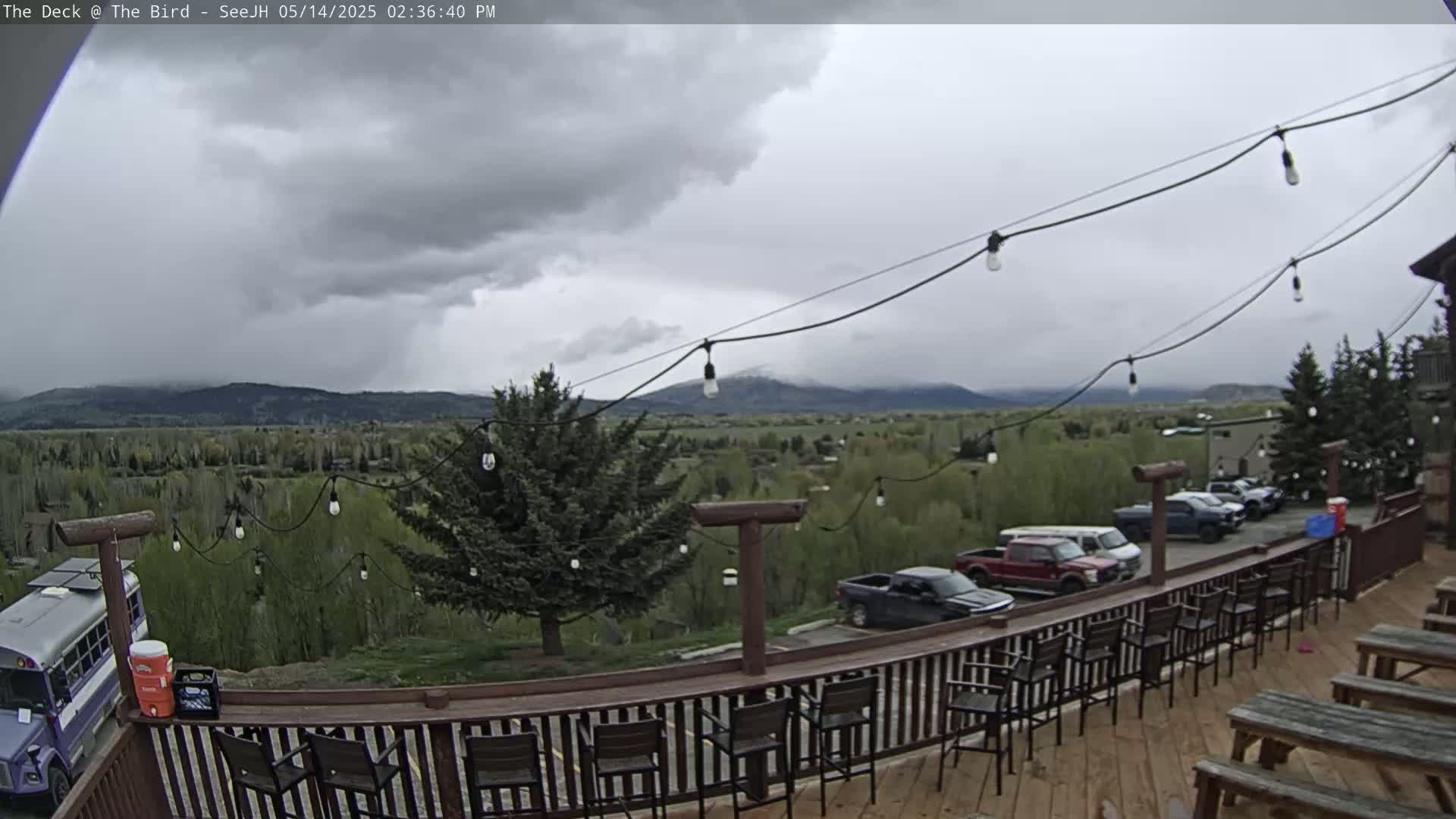 A deck with bar stools and picnic tables overlooks a valley of green trees and distant mountains under a cloudy sky.