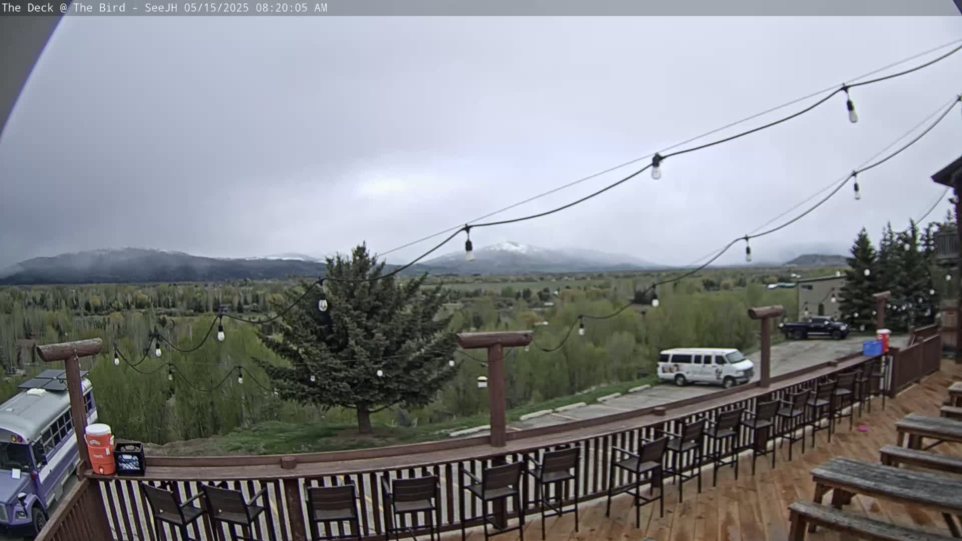 A deck with several empty chairs and picnic tables overlooks a valley of green trees and snow-capped mountains under an overcast sky.