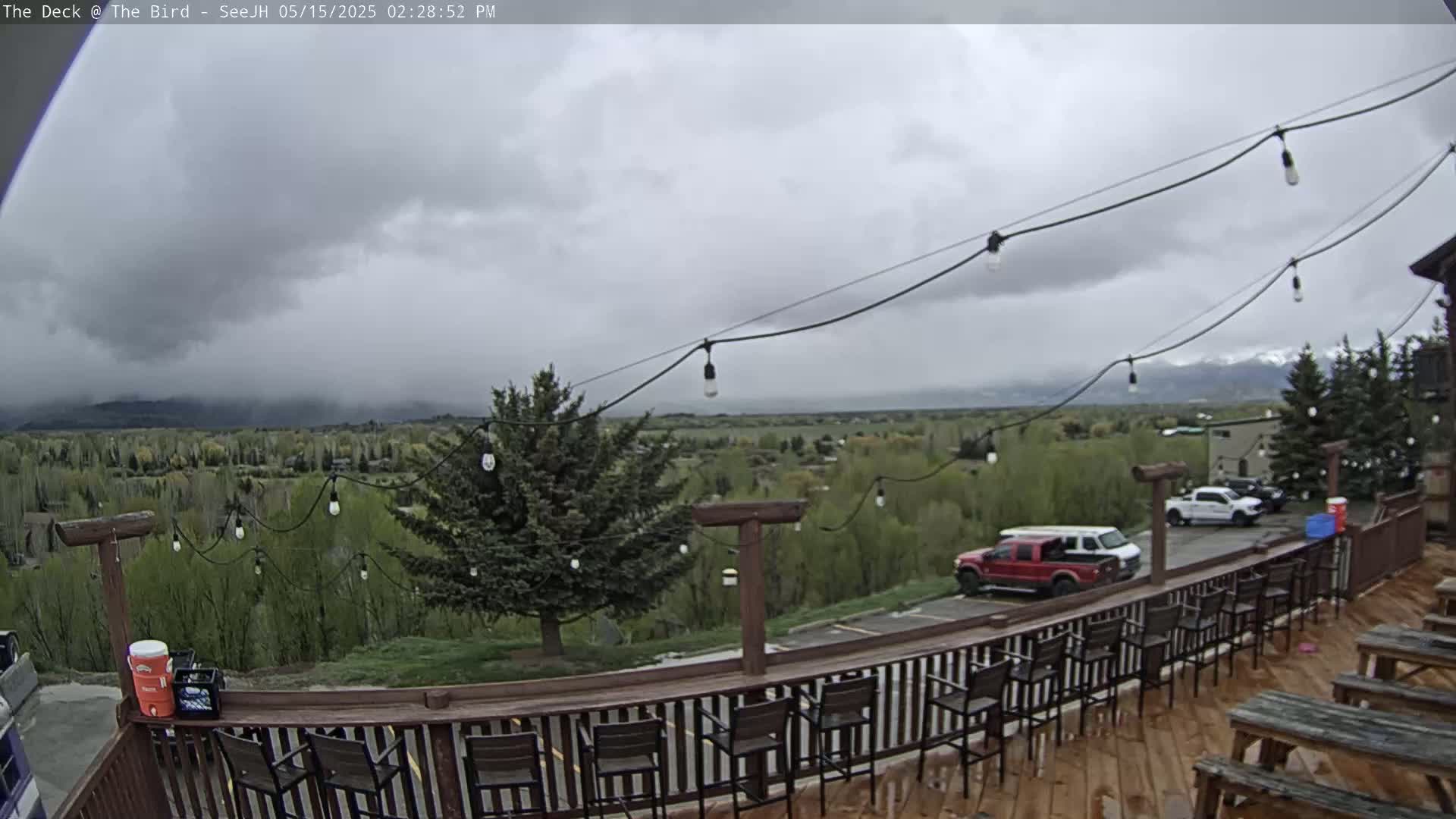 An outdoor deck with bar stools and picnic tables overlooks a valley of green trees and distant mountains under an overcast sky.