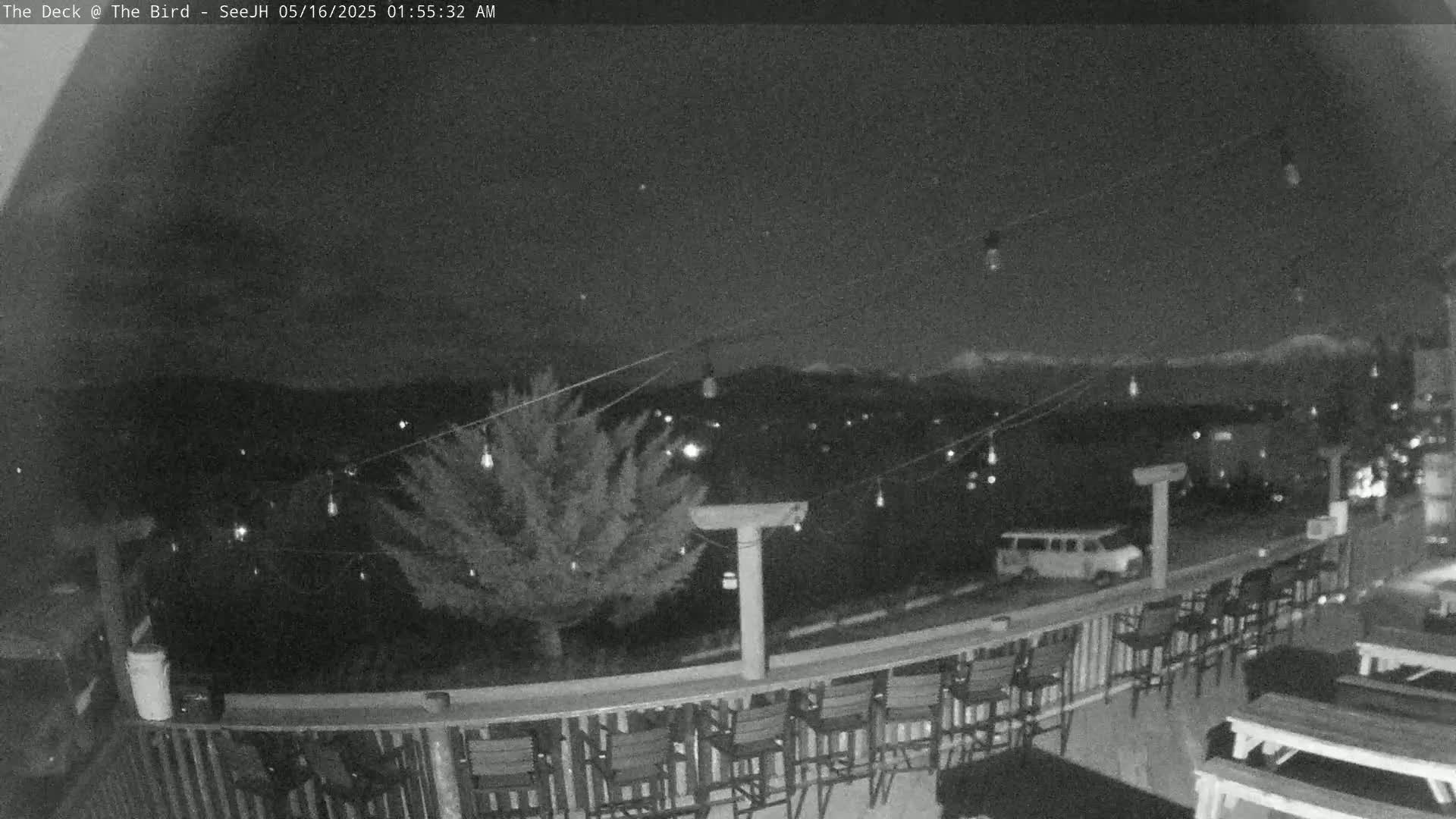 A nighttime view from a deck shows a row of empty bar stools, picnic tables, and a van parked beyond a railing overlooking a dimly lit town and distant mountains under a dark sky.