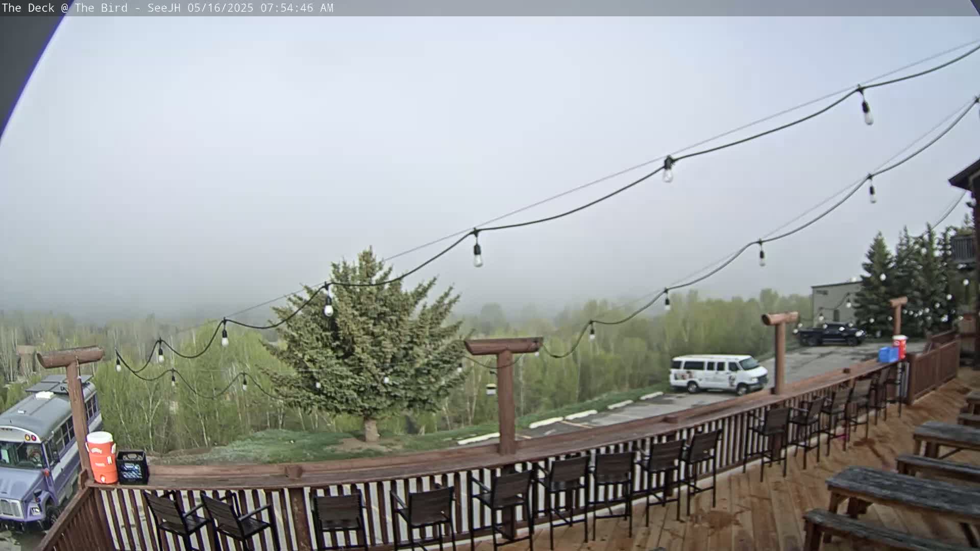 A foggy morning view from a wooden deck shows a row of empty bar stools, picnic tables, and string lights overlooking a parking lot with a bus and van, and a line of trees in the distance.