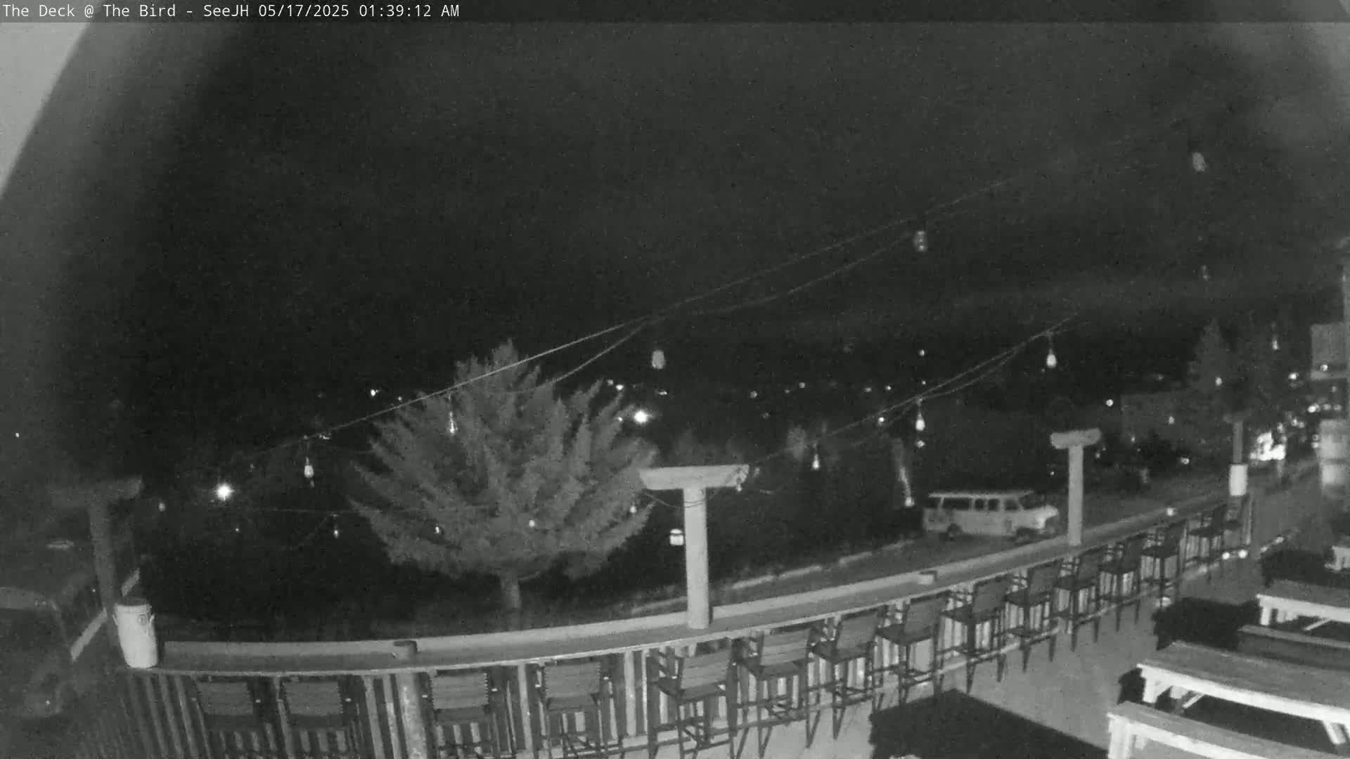 A nighttime view of an outdoor deck with string lights, several empty chairs and tables, and a vehicle parked nearby.