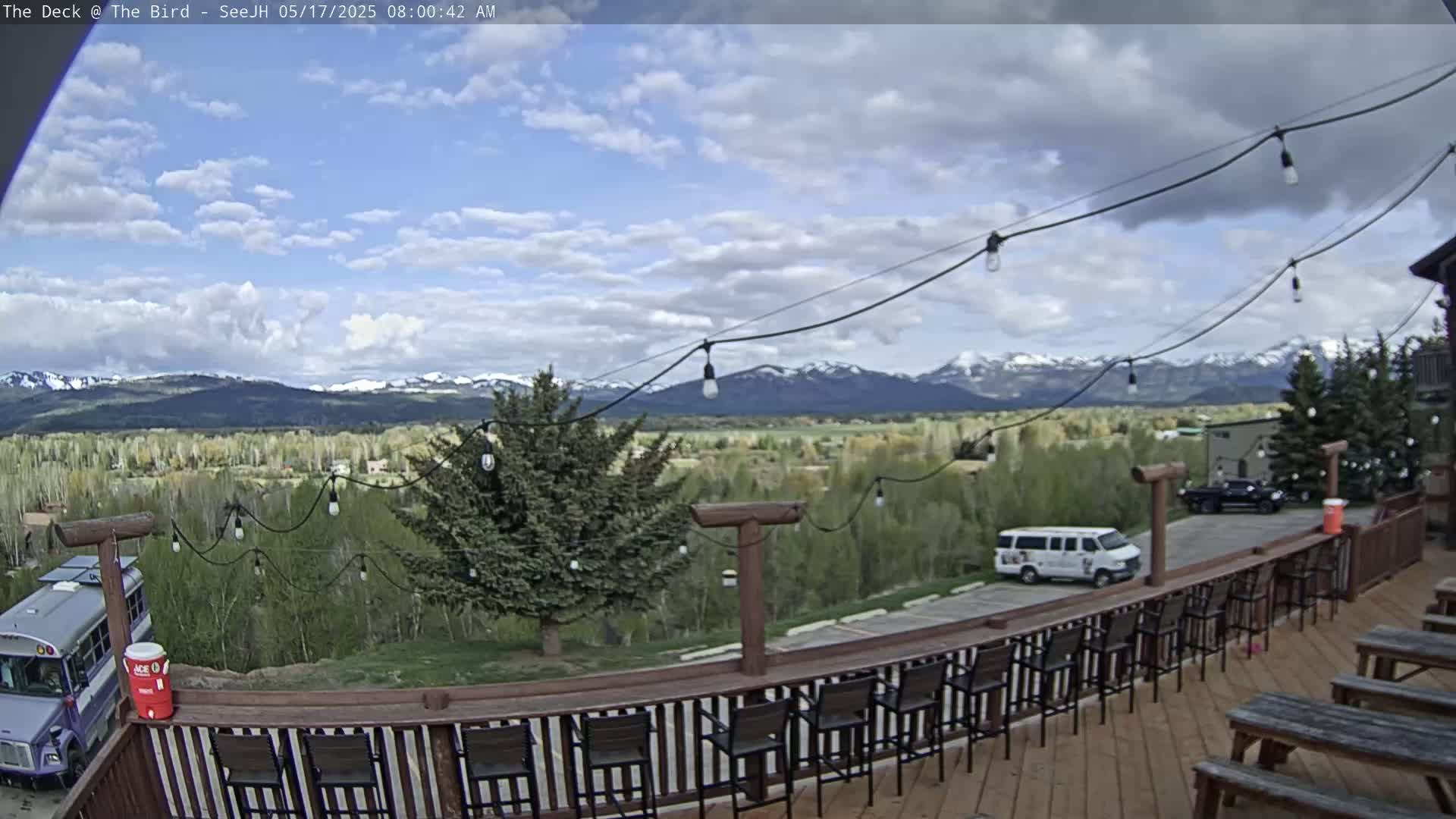 A partly cloudy outdoor scene shows a wooden deck with chairs and tables overlooking a valley, trees, and snow-capped mountains in the distance.