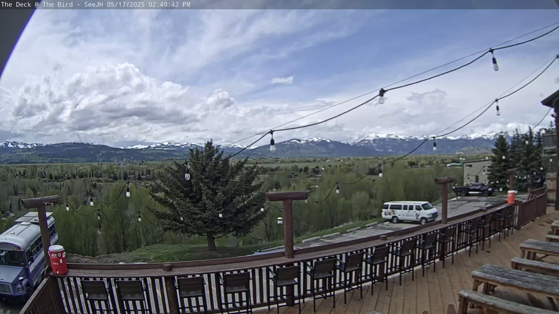 A partly cloudy day reveals a mountainous landscape viewed from a wooden deck with string lights and several empty chairs and picnic tables.