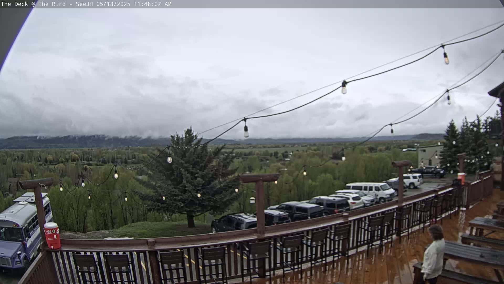 An outdoor deck with string lights overlooks a valley and mountains under a cloudy sky, with several vehicles parked nearby and a person standing on the deck.