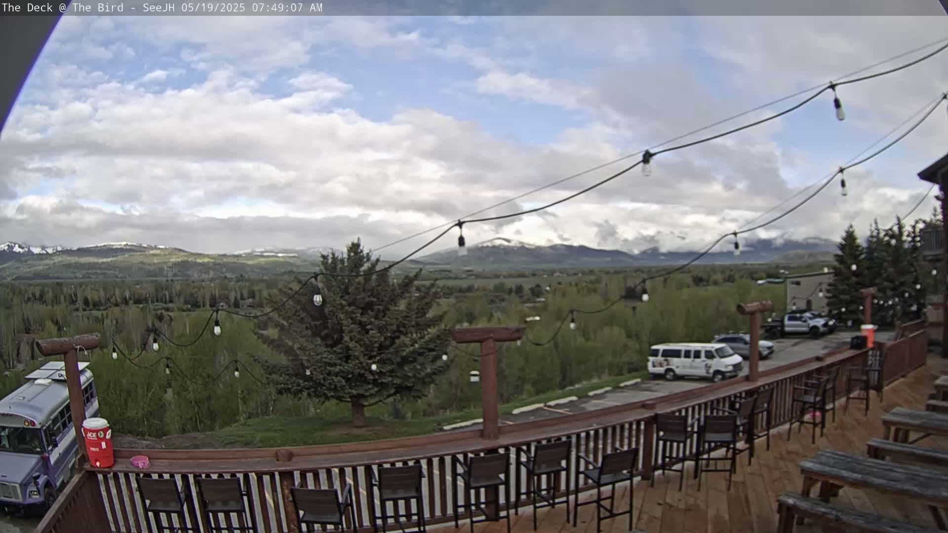 A partly cloudy outdoor scene shows a deck with empty chairs and tables overlooking a valley with distant mountains, a school bus, and a few vehicles parked nearby.