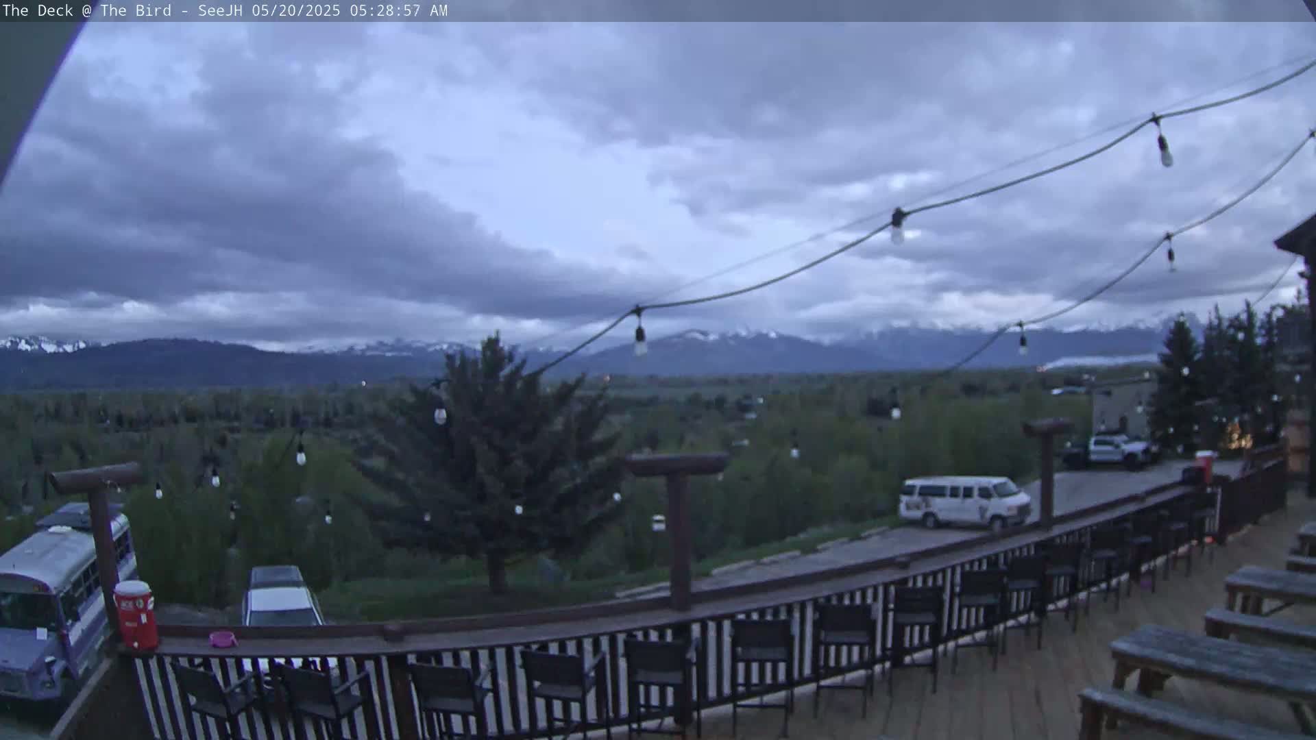 An outdoor deck with string lights overlooks a valley, mountains, and parked vehicles under a cloudy sky.