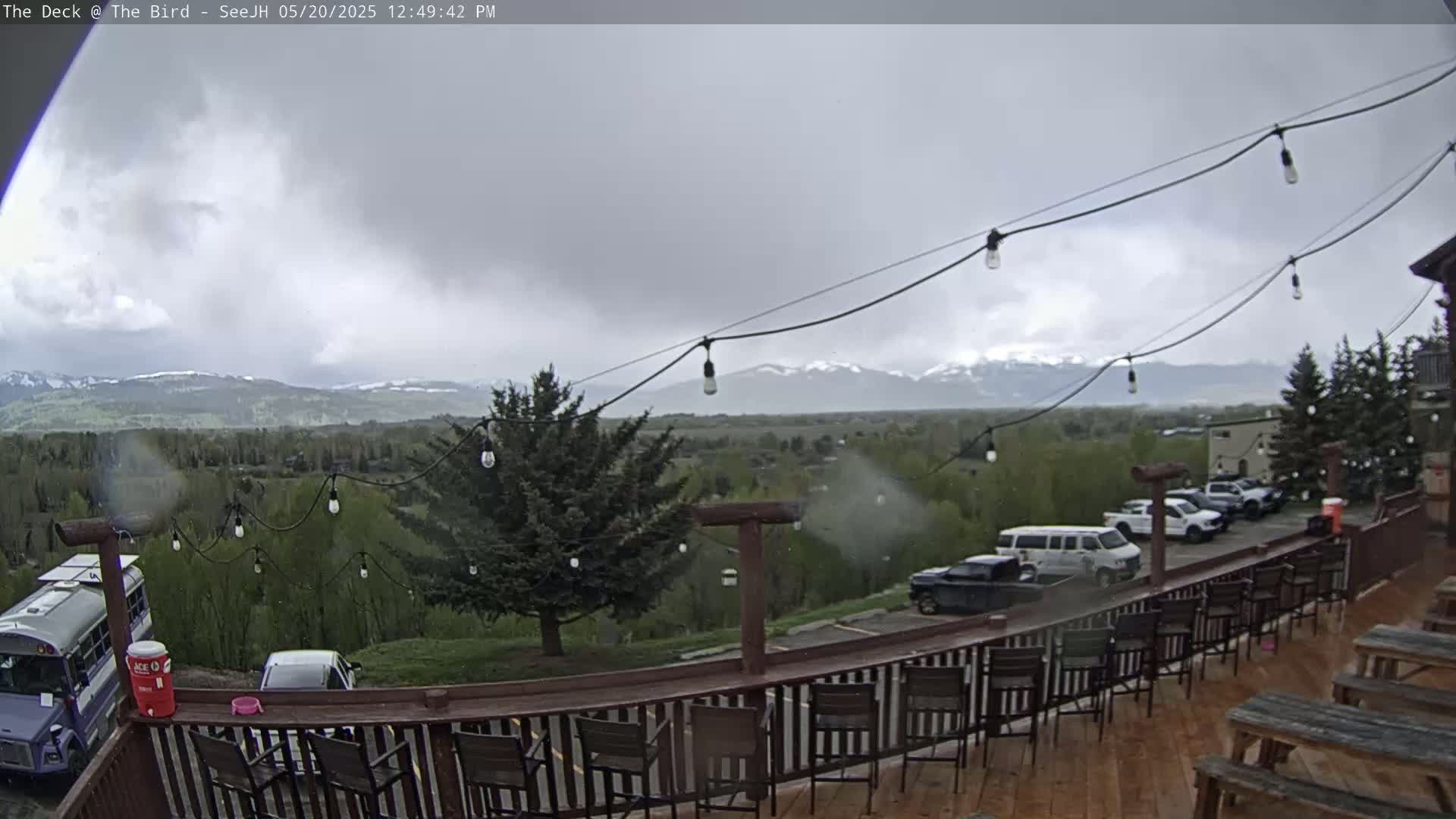 A deck with chairs and picnic tables overlooks a valley of green trees and snow-capped mountains under a cloudy, possibly rainy sky.