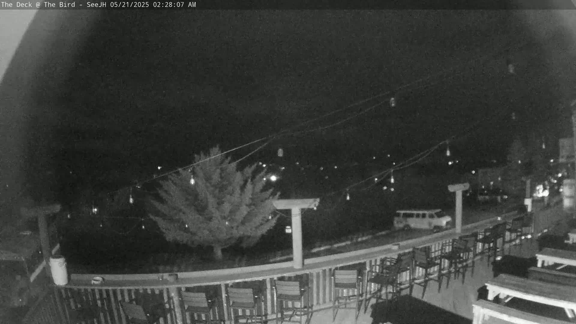 A nighttime view of an outdoor deck with string lights, chairs, and tables, overlooking a tree and a parking lot.