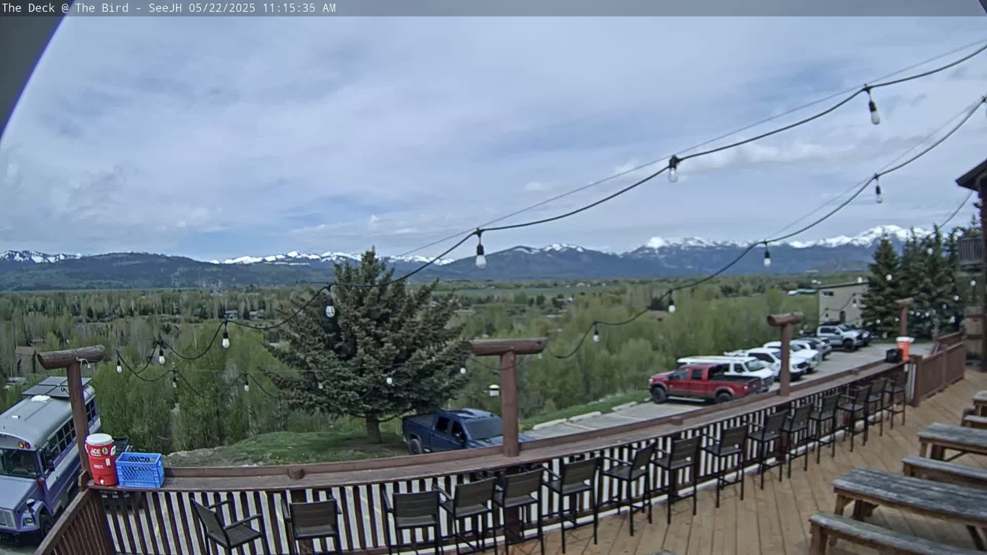 A partly cloudy outdoor deck scene features a mountain range in the background, several vehicles parked nearby, and empty bar stools lining a wooden railing.