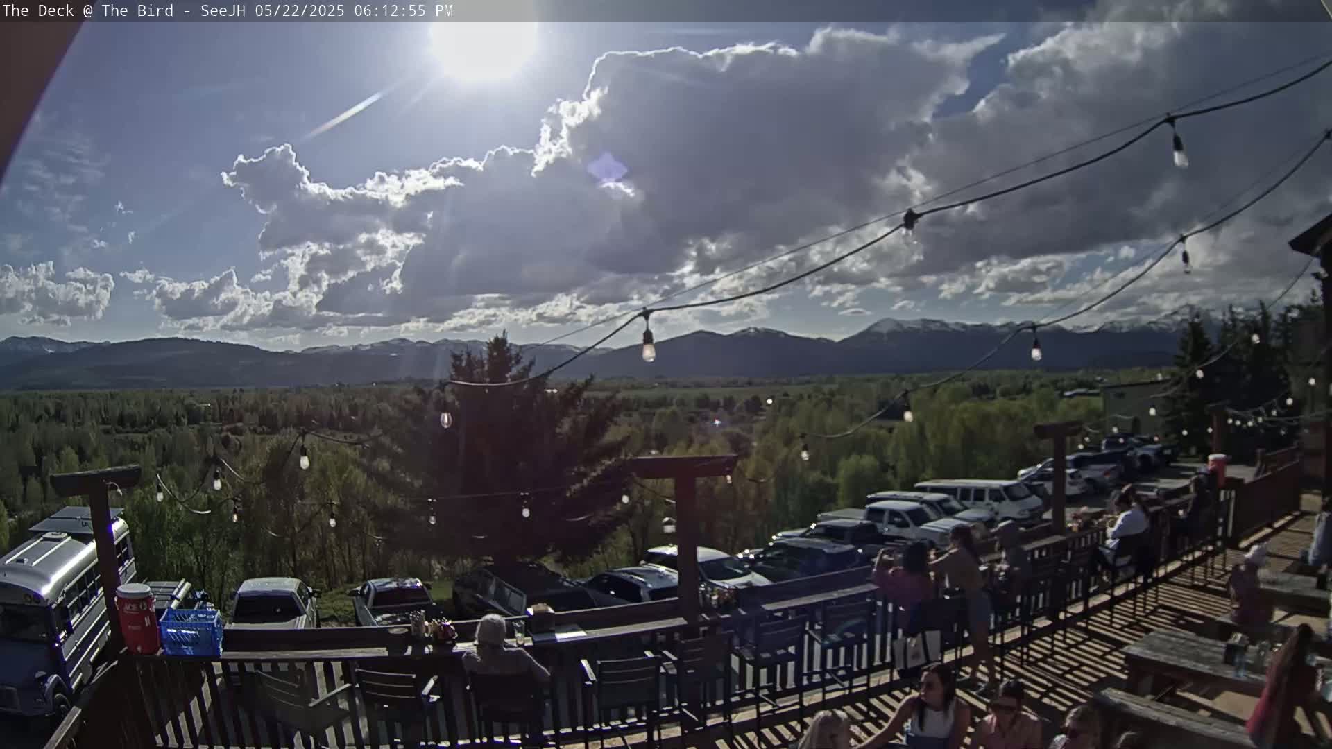 A partly cloudy outdoor deck scene features a mountain range in the background, string lights overhead, and numerous people sitting at tables and a bar.