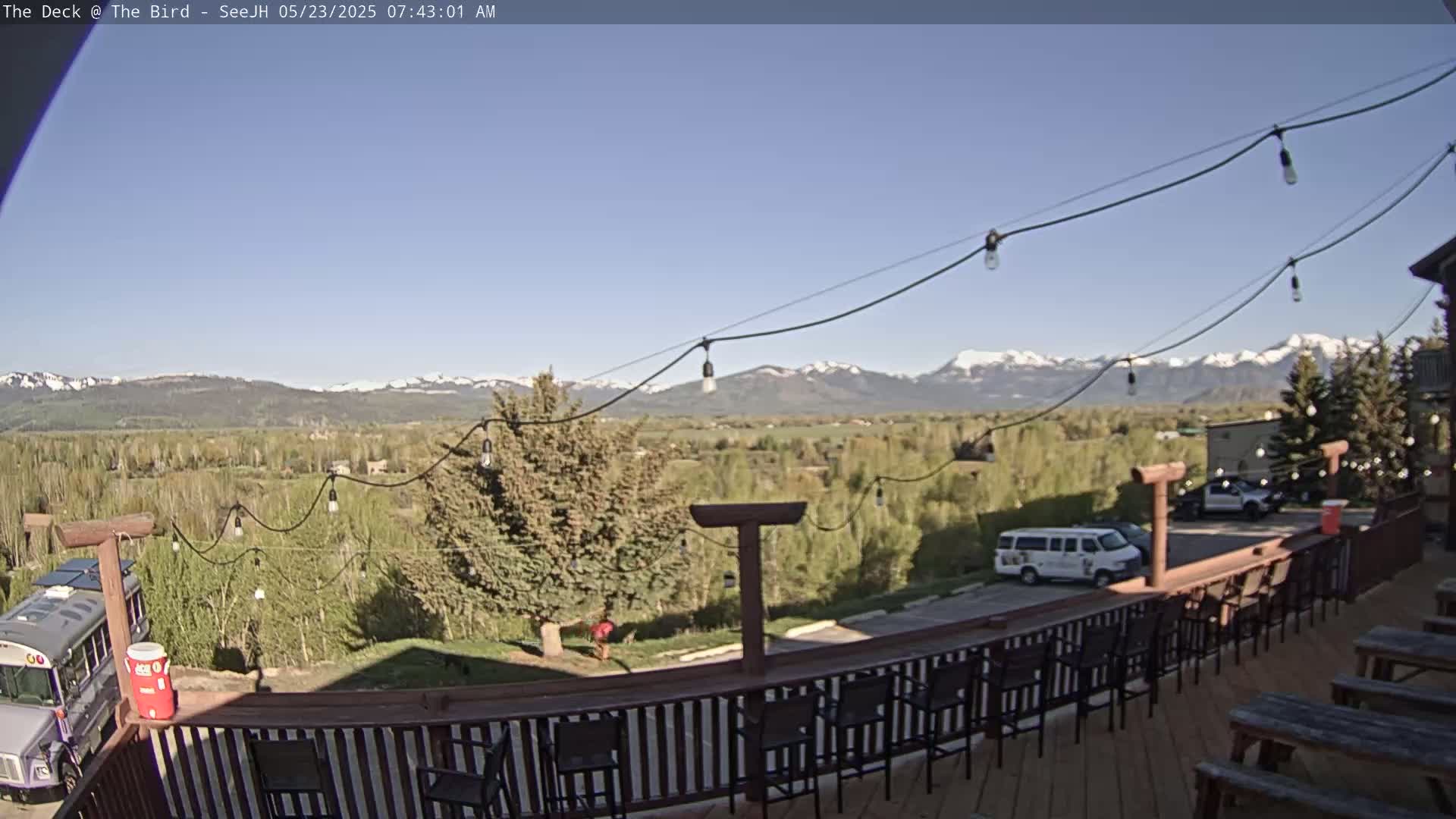 A deck with string lights overlooks a valley and snow-capped mountains under a clear blue sky.