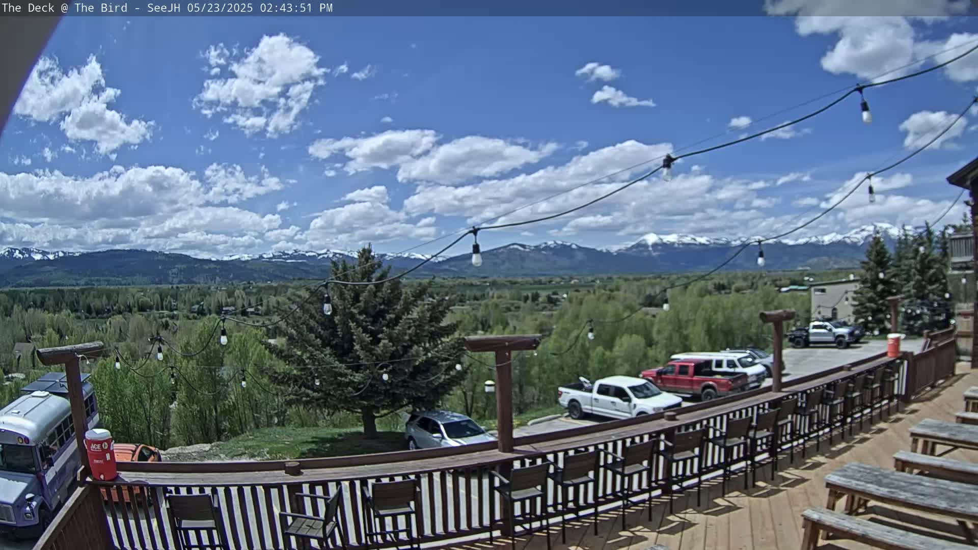 A deck with a bar and seating overlooks a valley of green trees and distant snow-capped mountains under a partly cloudy blue sky.