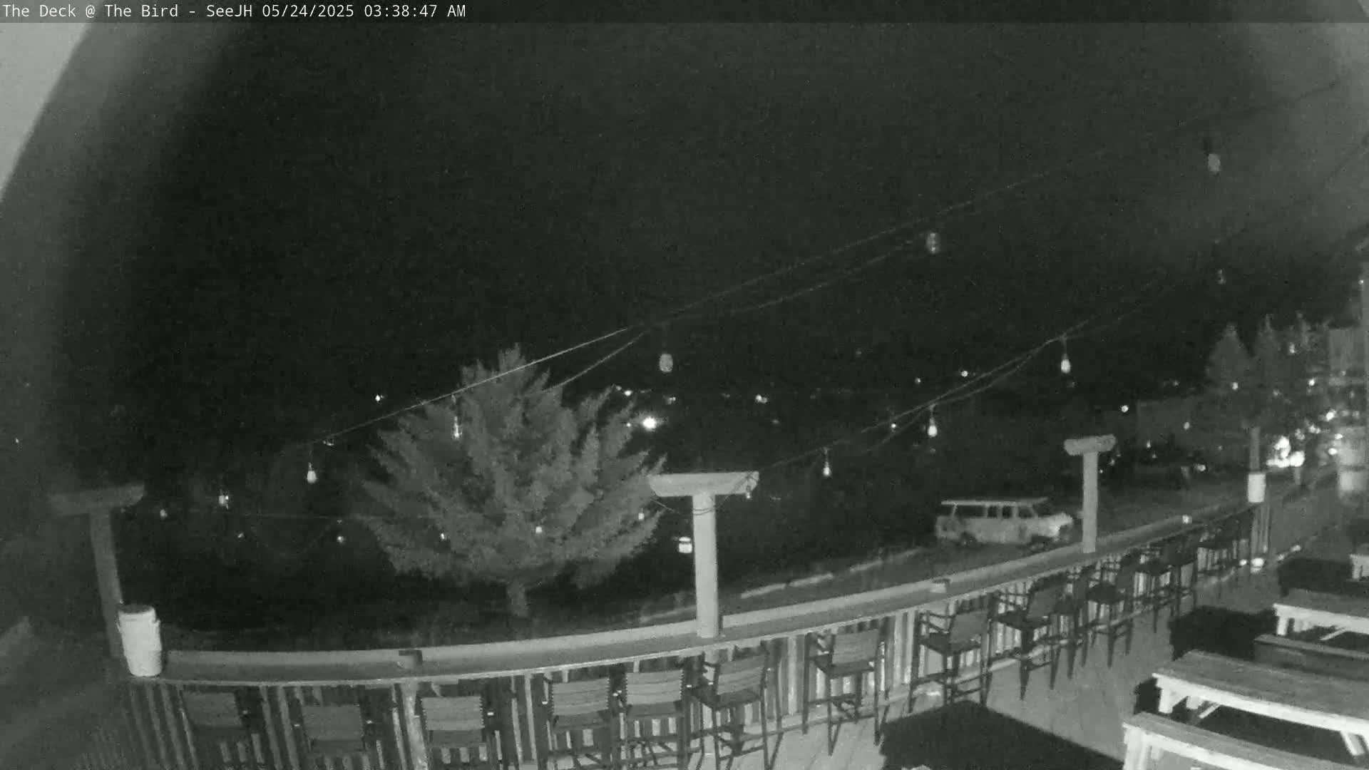 A nighttime view of an outdoor deck with string lights, chairs, and tables, overlooking a parking lot with a van.