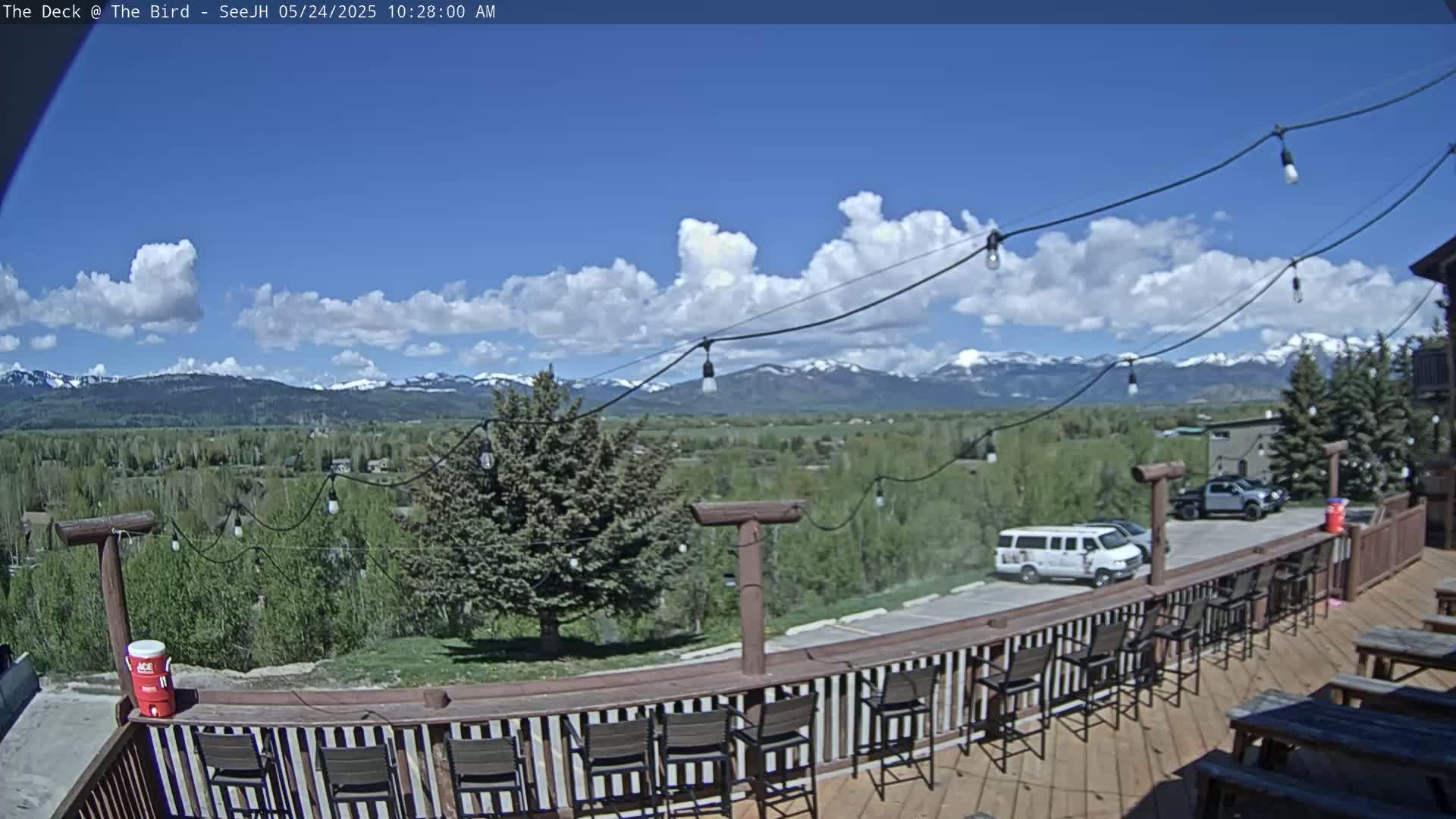 A deck with empty chairs and tables overlooks a valley of green trees and distant snow-capped mountains under a partly cloudy blue sky.
