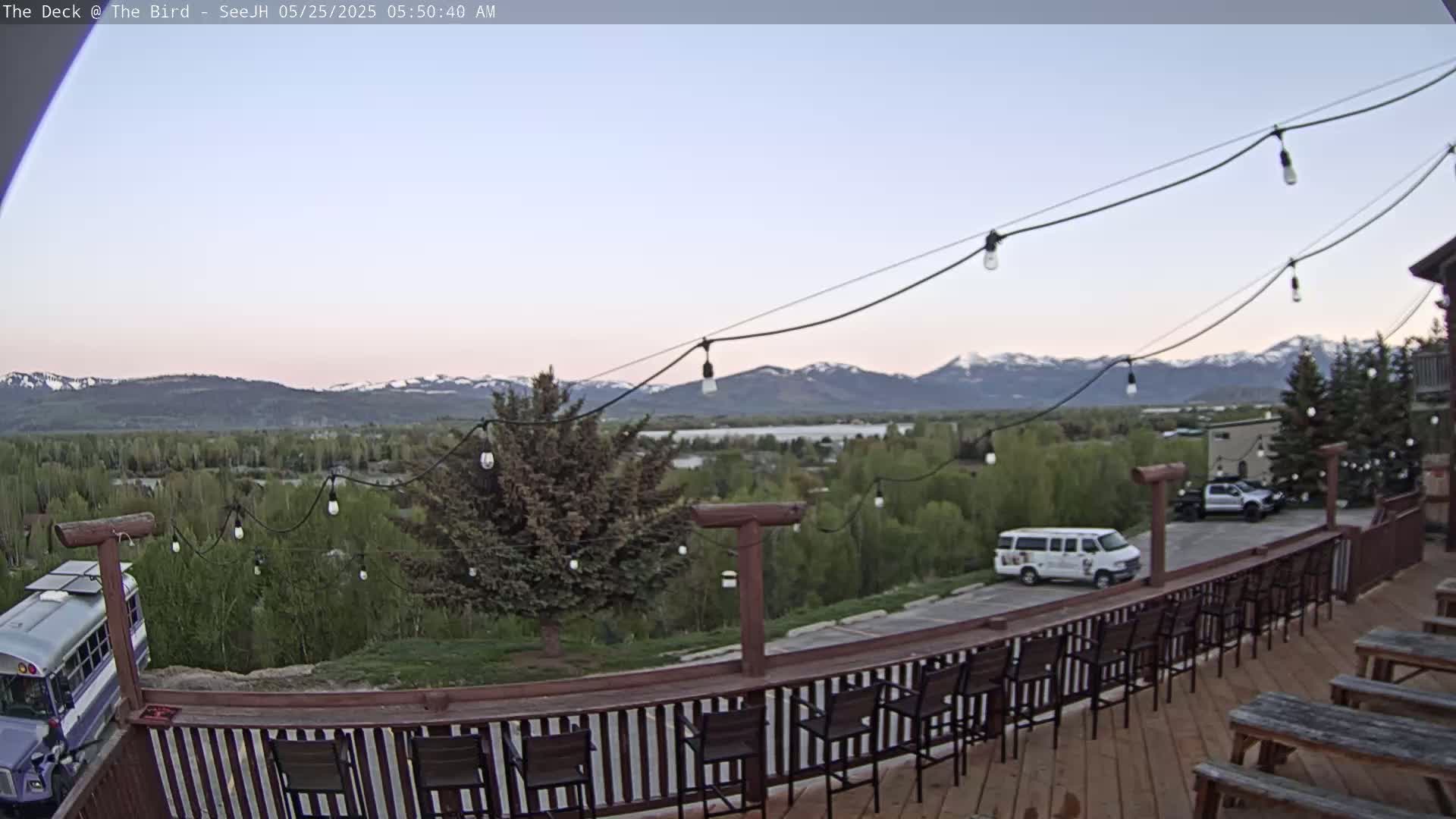 An outdoor deck with string lights and empty bar stools overlooks a valley and distant snow-capped mountains under a clear, twilight sky.