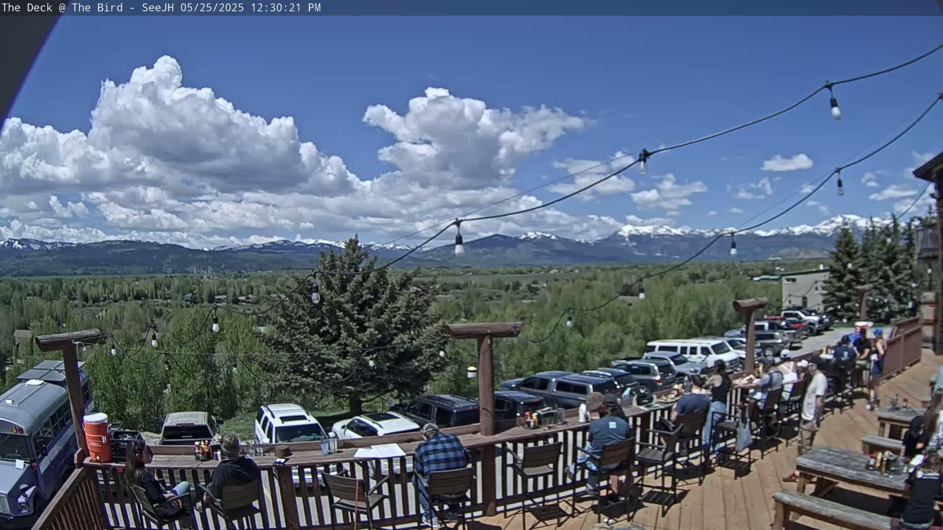 A sunny outdoor patio with people seated at tables overlooking a valley and snow-capped mountains under a partly cloudy sky.