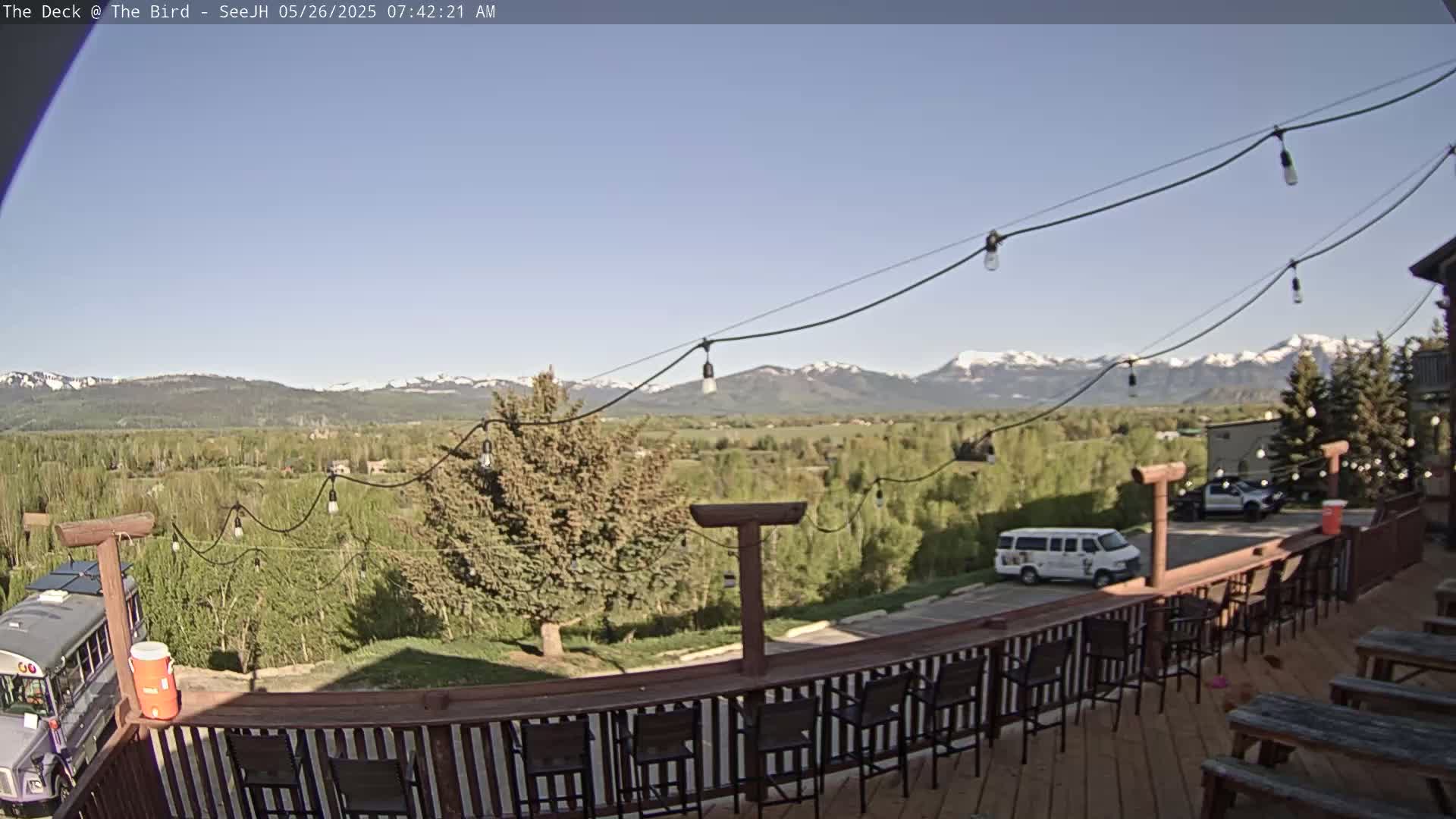 A wooden deck with outdoor seating overlooks a valley of green trees and distant snow-capped mountains under a clear blue sky.