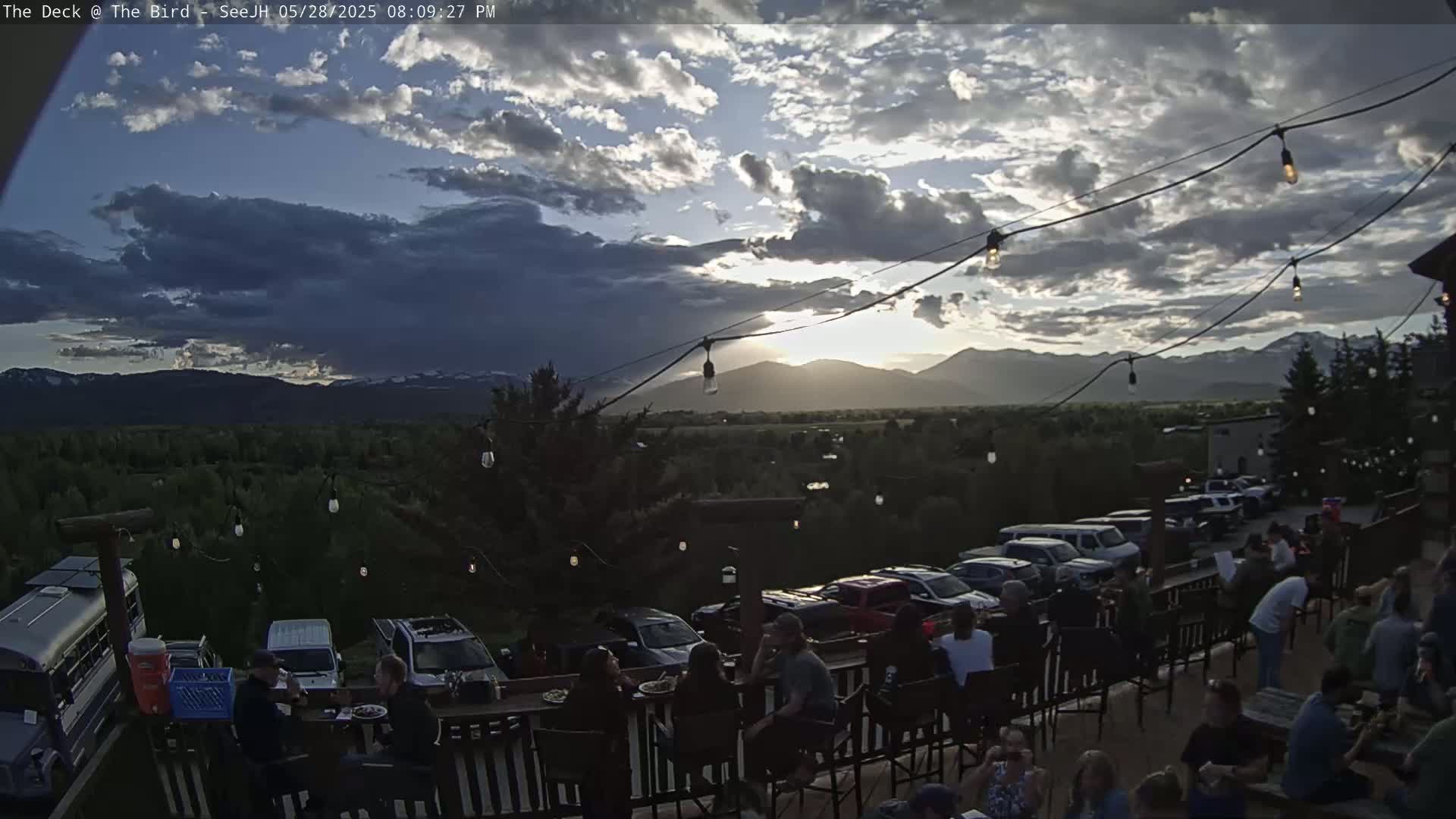 An outdoor patio with people seated at tables overlooks a valley and mountains at sunset under a partly cloudy sky.
