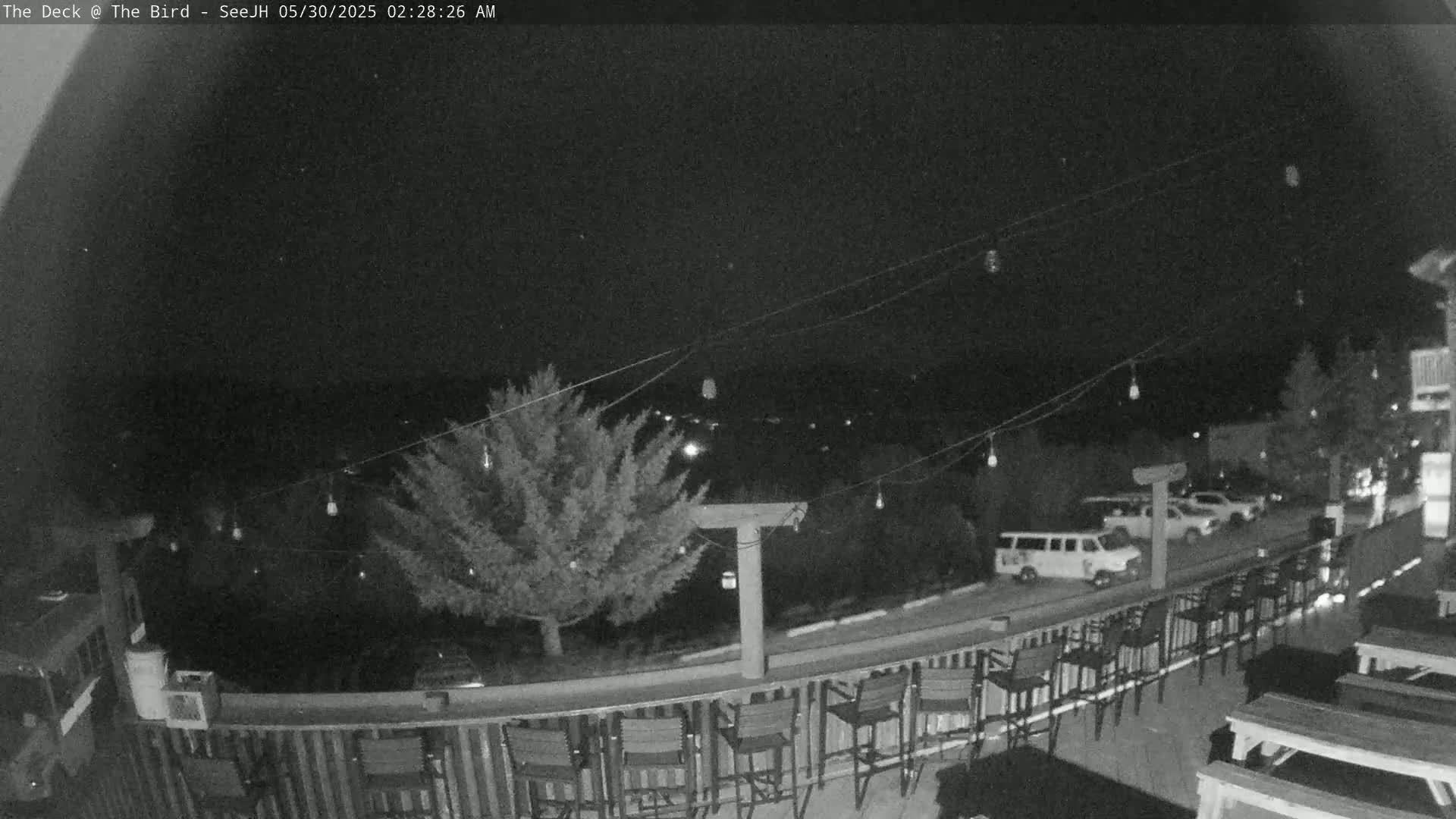 A nighttime view of an outdoor deck with several empty chairs and picnic tables, string lights overhead, and a few vehicles parked nearby.