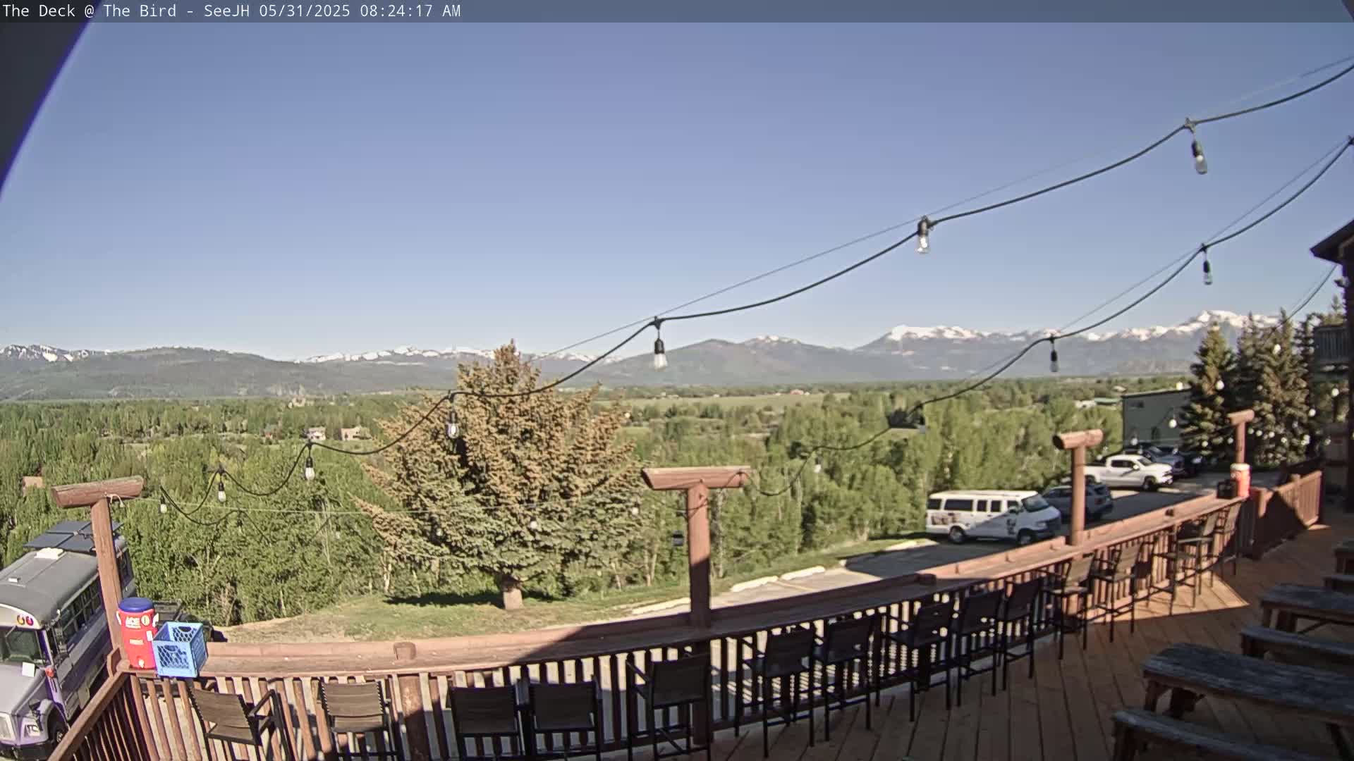 A wooden deck with chairs and tables overlooks a valley of green trees and distant snow-capped mountains under a clear blue sky.