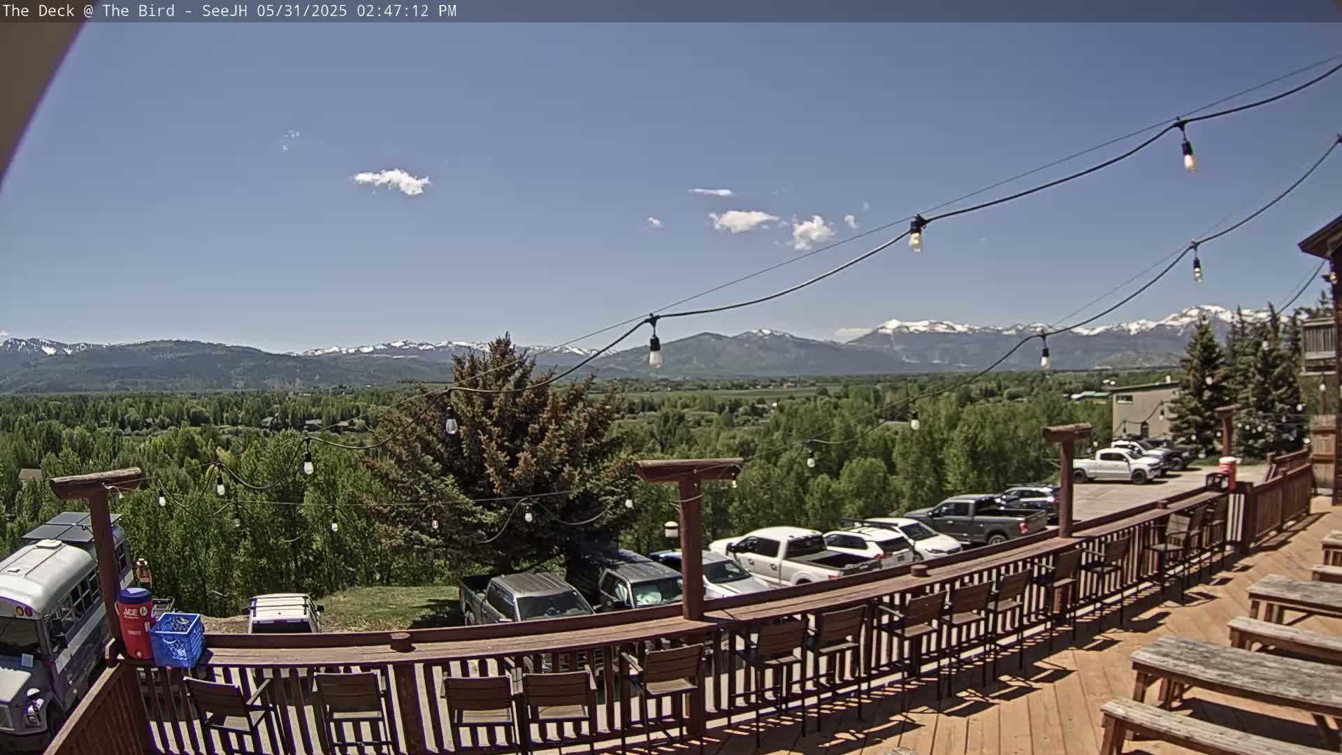 A deck with bar stools and picnic tables overlooks a valley of green trees and distant snow-capped mountains under a mostly clear, sunny sky.