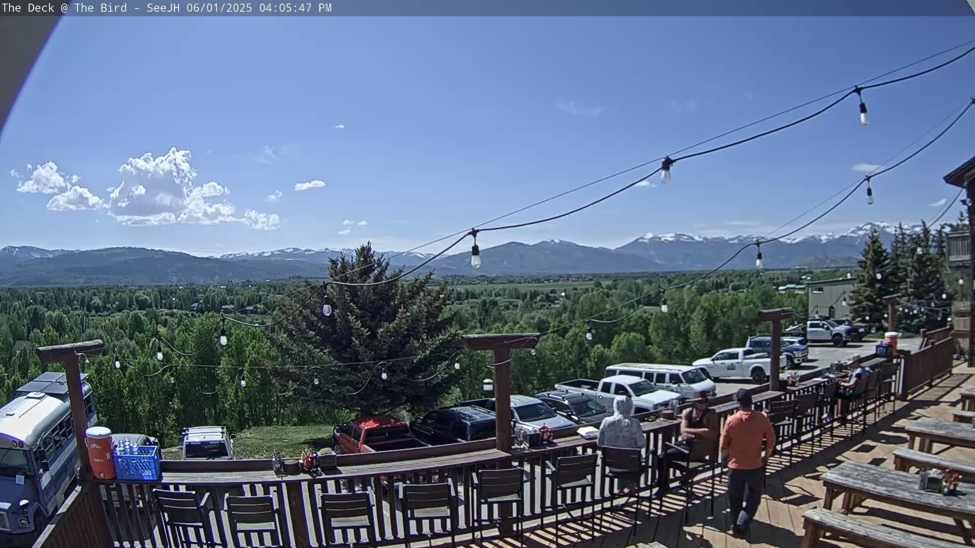 A sunny outdoor deck overlooks a valley and snow-capped mountains, with several people seated at a bar and tables.