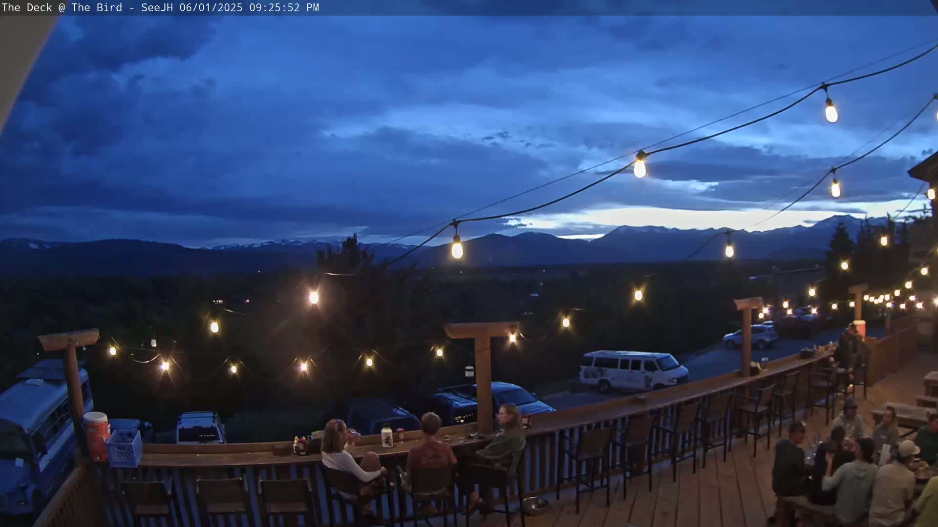An outdoor deck at dusk, lit by string lights, overlooks a mountain range under a partly cloudy sky.