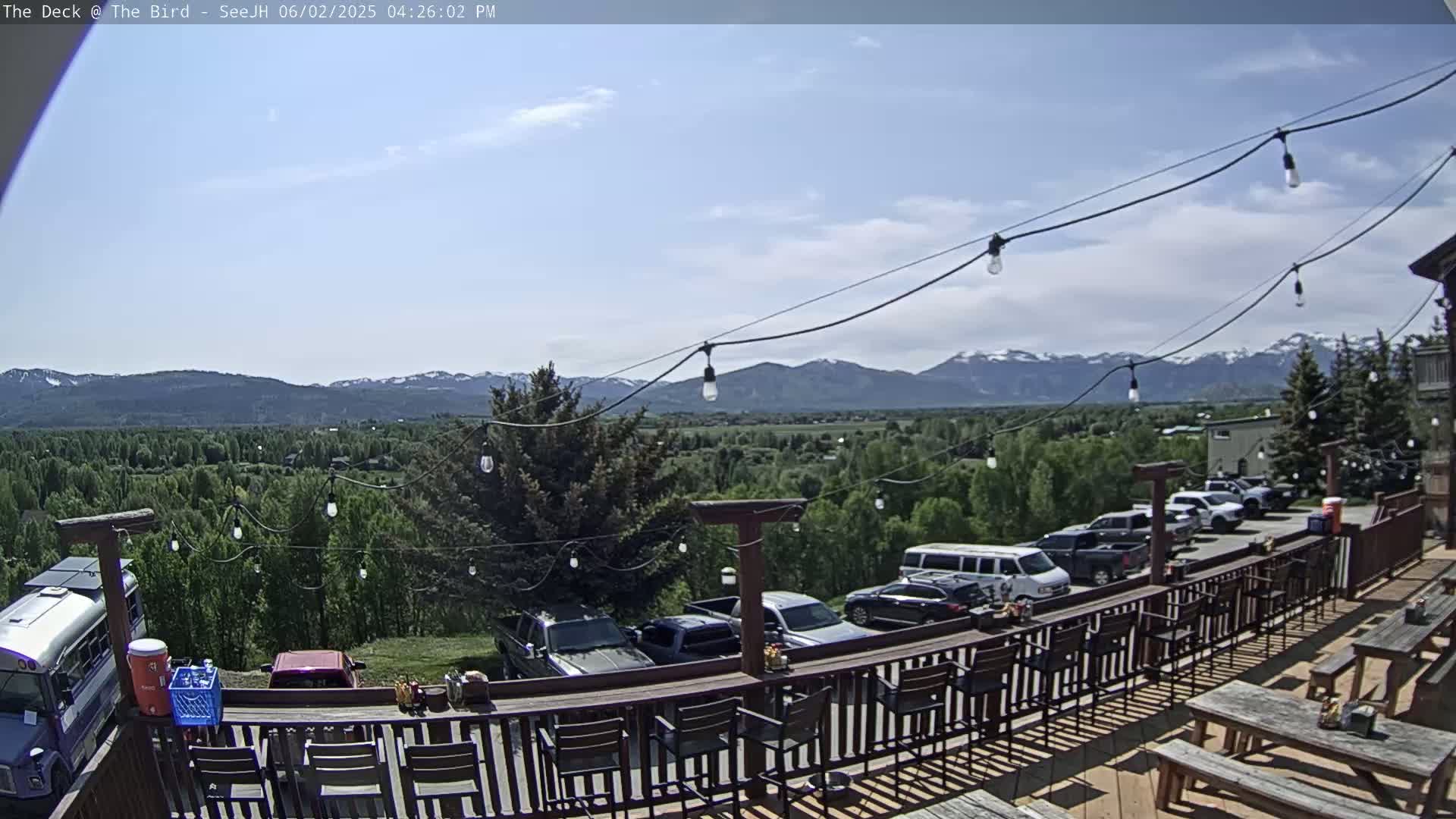 A sunny outdoor deck with a bar and picnic tables overlooks a valley and distant mountains under a partly cloudy sky.