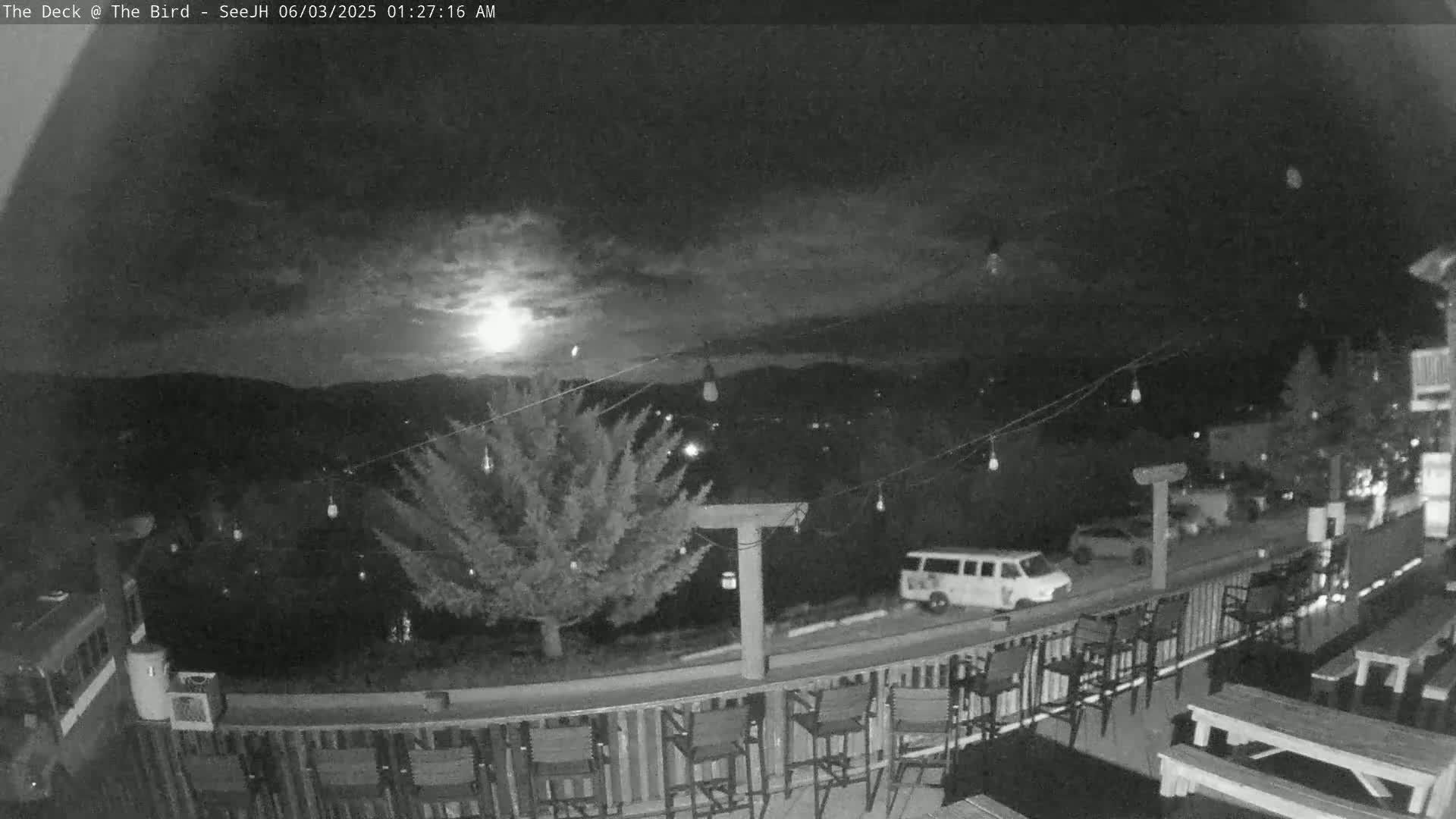 A nighttime view from a deck shows a partly cloudy sky with a bright moon, string lights, a tree, a van parked on a street, and empty chairs and picnic tables on the deck.