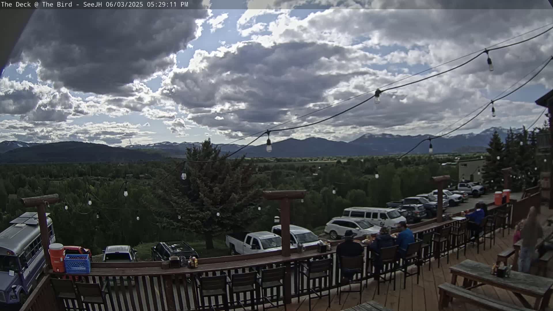 A partly cloudy outdoor deck scene overlooking a valley with mountains in the distance, showing people seated at a bar, cars parked nearby, and string lights overhead.