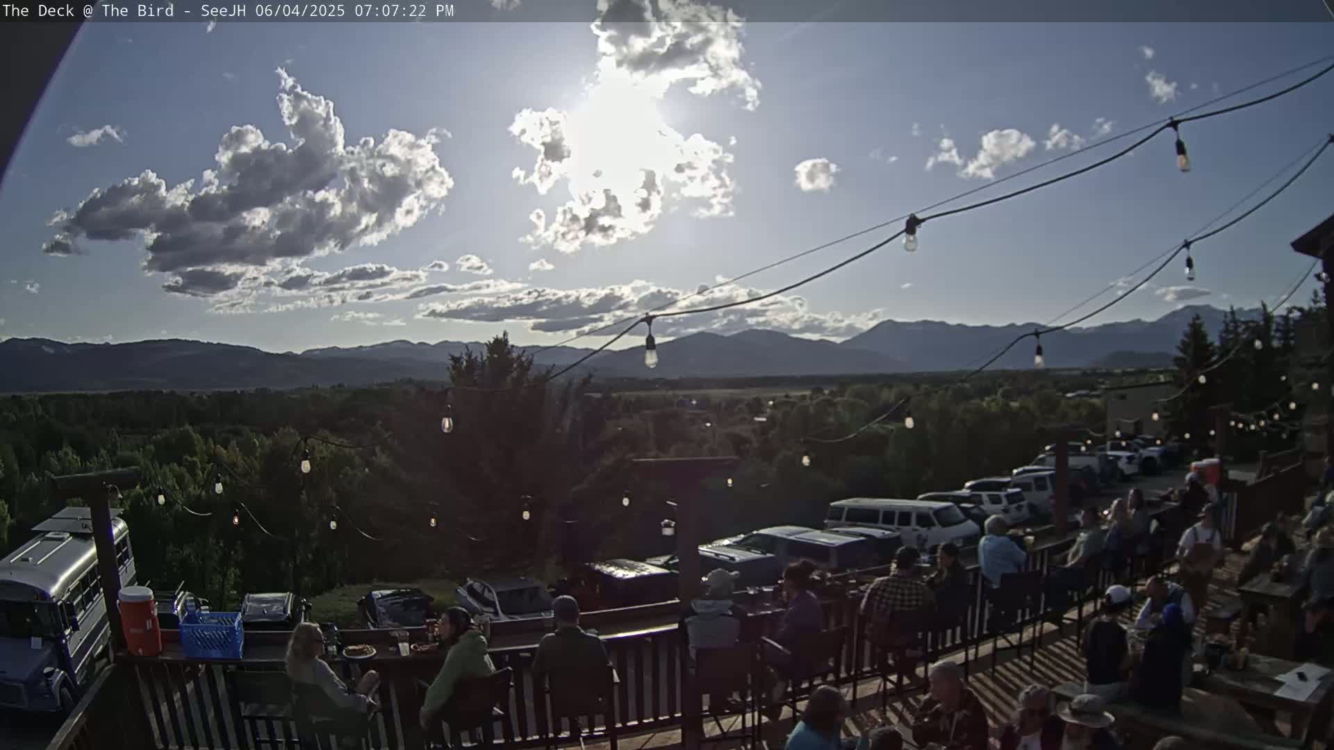 A partly sunny outdoor deck scene shows people seated at tables overlooking a mountain range and a valley of trees, with string lights overhead.