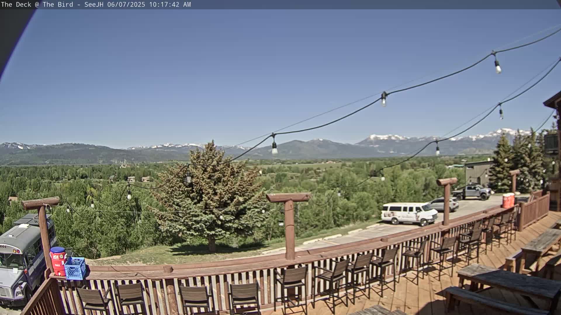 A deck with empty bar stools and picnic tables overlooks a valley of green trees and distant snow-capped mountains under a clear blue sky.