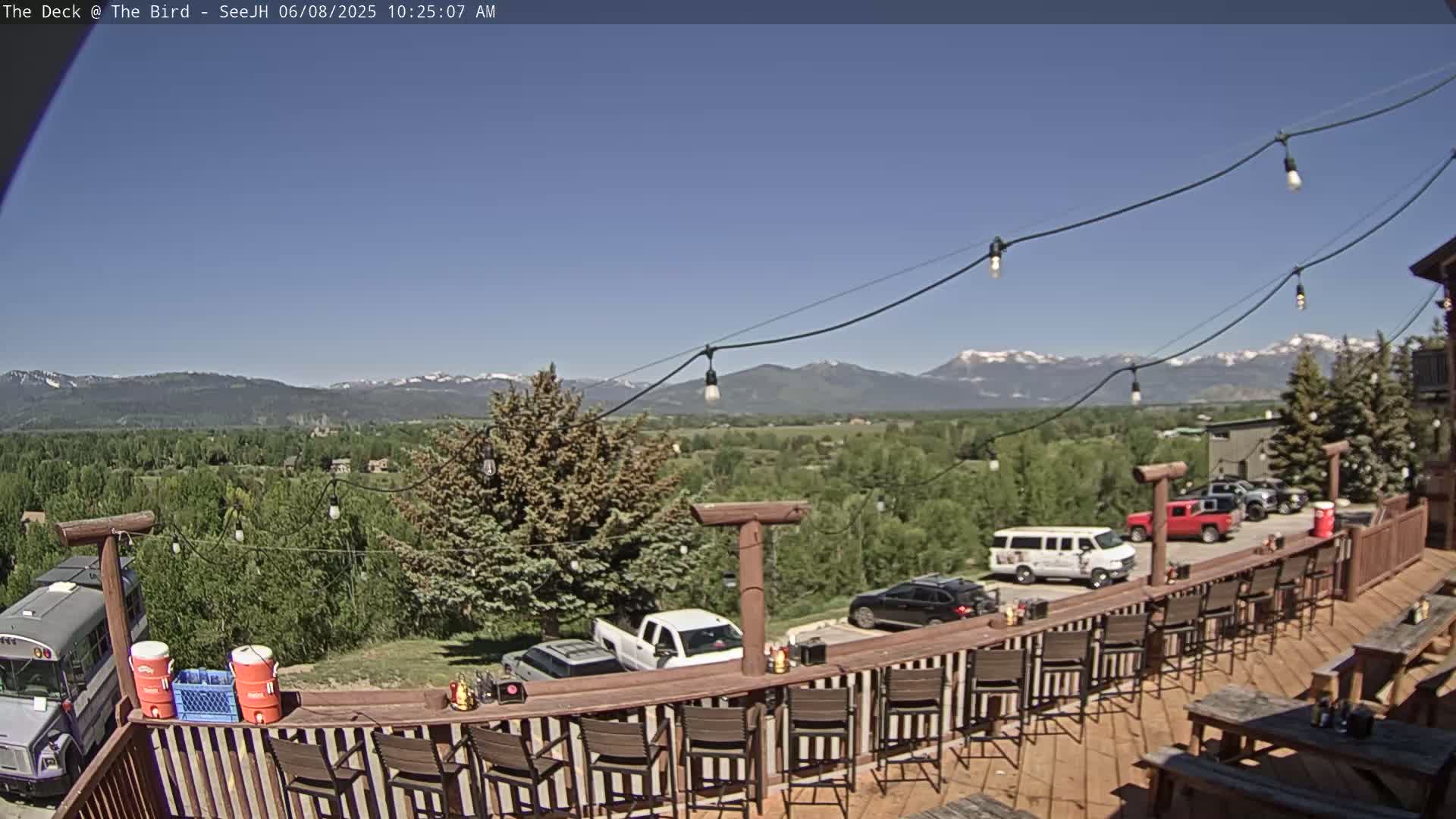 A deck with a bar and chairs overlooks a valley of green trees and distant snow-capped mountains under a clear blue sky.