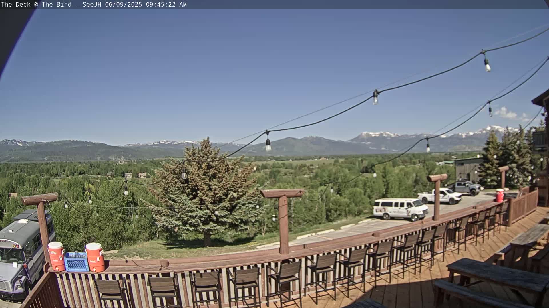 A deck with bar stools overlooks a green valley and snow-capped mountains under a clear blue sky.