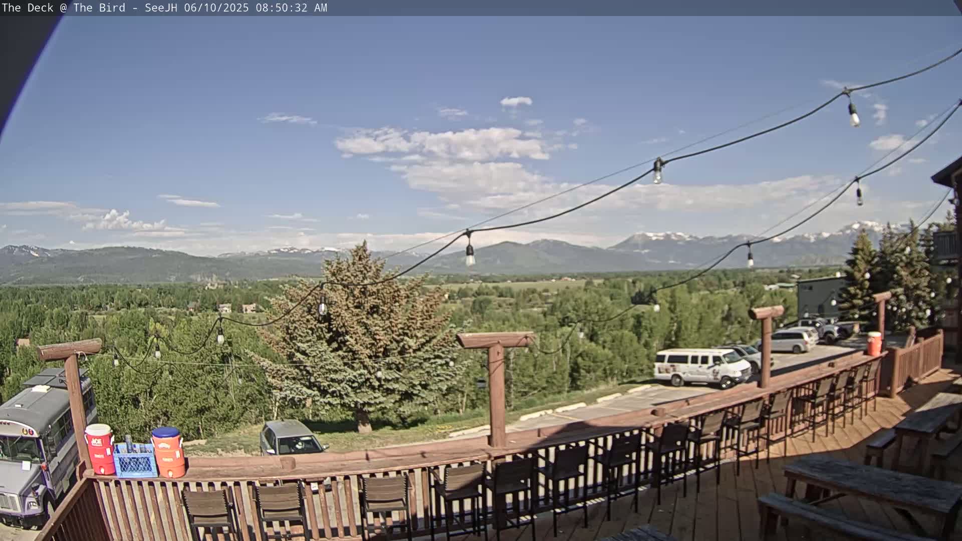 An outdoor deck with a wooden railing and barstools overlooks a valley, mountains, and a partly cloudy sky.