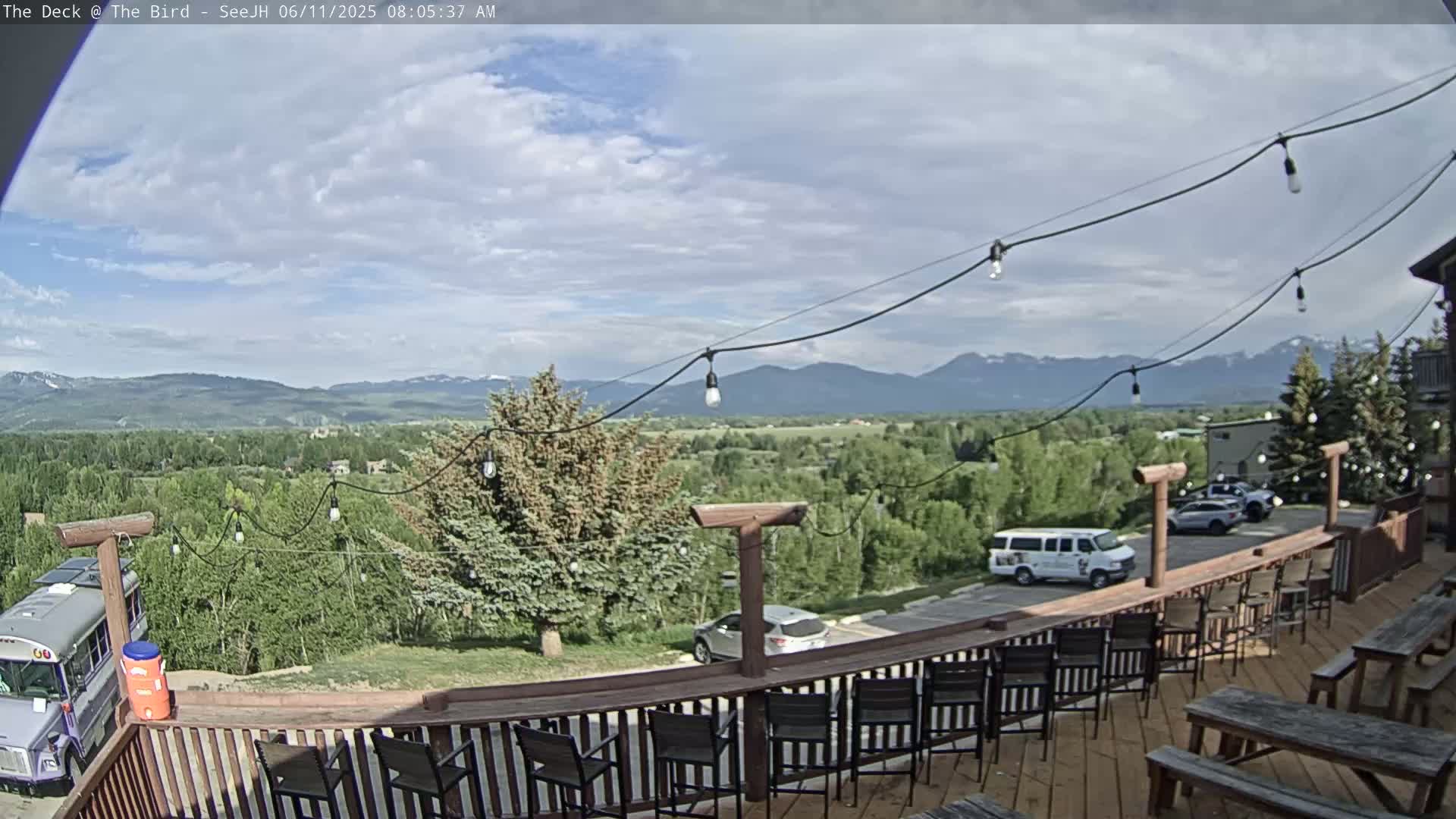A partly cloudy day reveals a mountain range in the distance, viewed from a deck furnished with chairs and picnic tables, string lights overhead.
