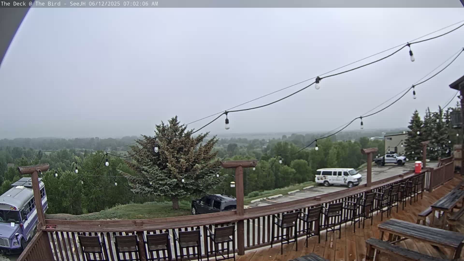 A hazy, overcast outdoor scene shows a wooden deck with chairs and tables overlooking a parking lot and a distant, tree-covered landscape.